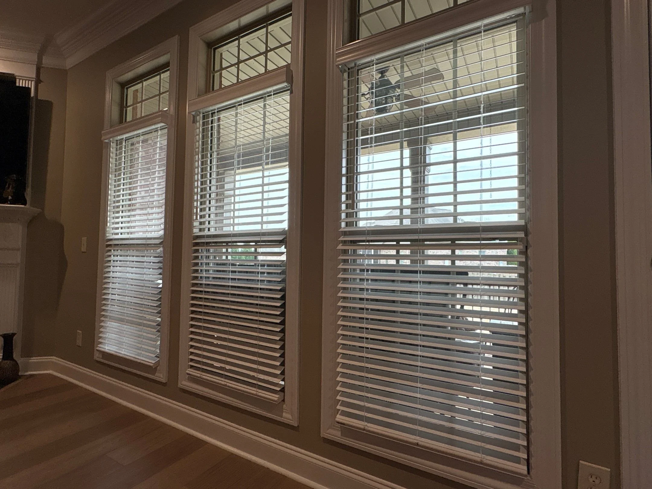 A set of indoor windows with white blinds, three of which are partially open. Outside, there is a porch with a ceiling fan and a hanging light.