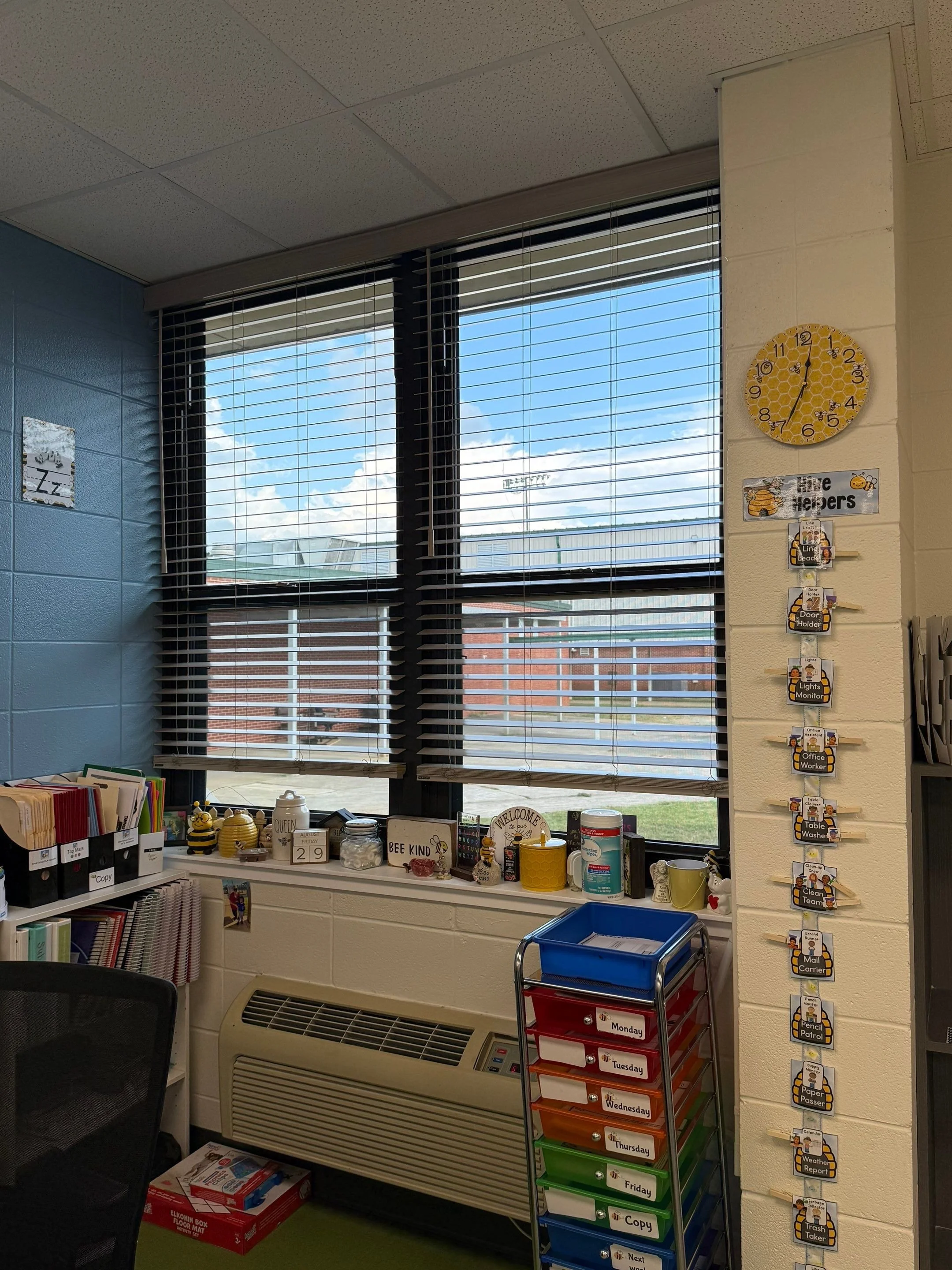 A classroom window with blue blinds showing a blue sky with white clouds outside. The classroom wall has a yellow clock, a sign that says 'Hive Helpers' with bee illustrations, and a vertical 'Tasks' chart with various classroom chores listed.