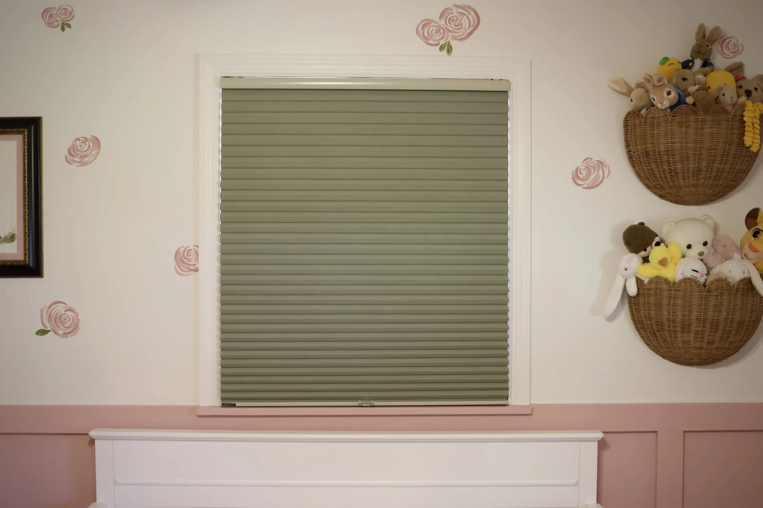 A window with closed green blinds, surrounded by pink wall with rose patterns, and decorated with two wicker baskets filled with stuffed animals and plush toys.