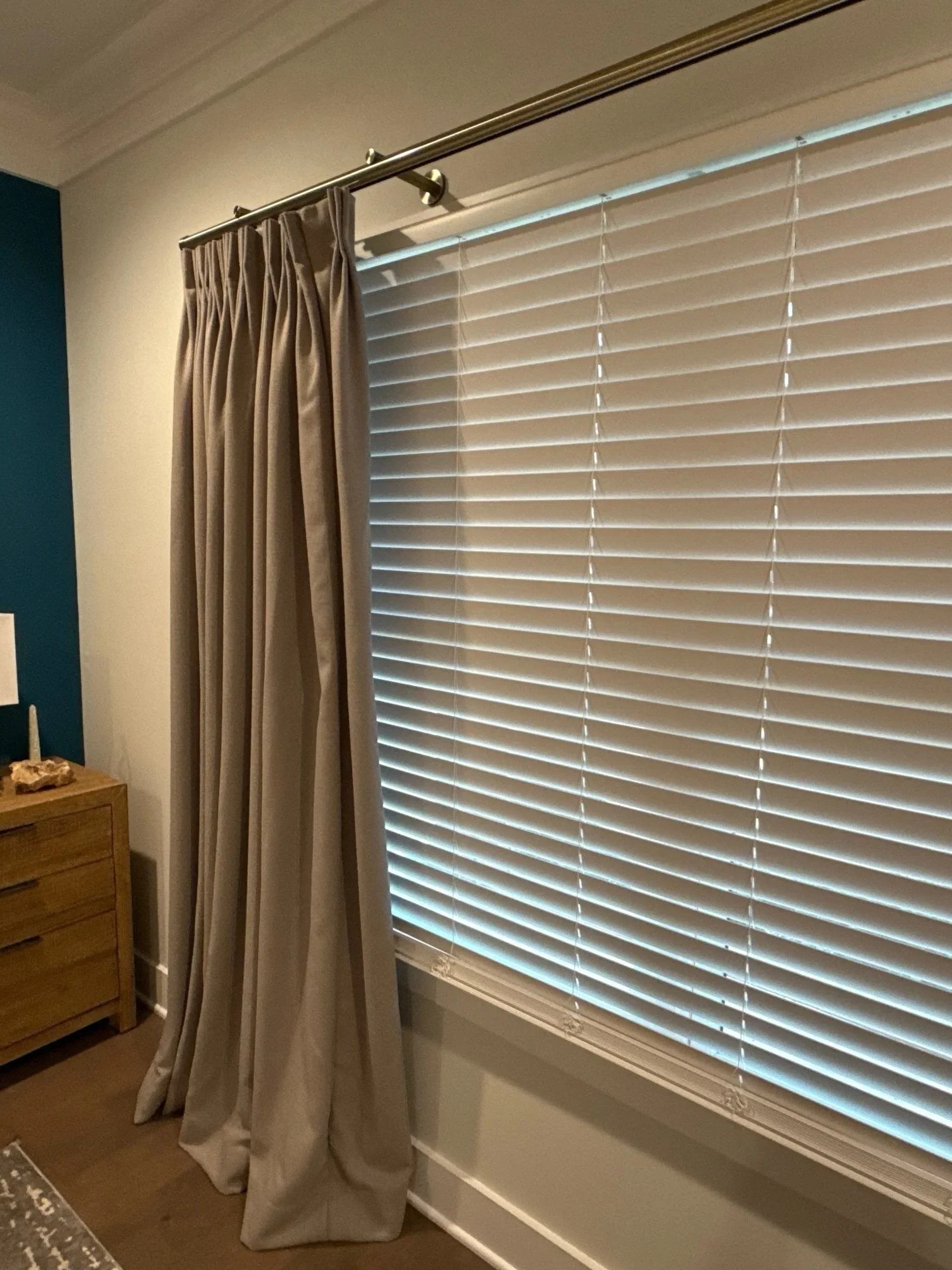 A window with closed blinds and beige curtains pulled back on a curtain rod. Part of a wooden dresser is visible on the left side of the image.