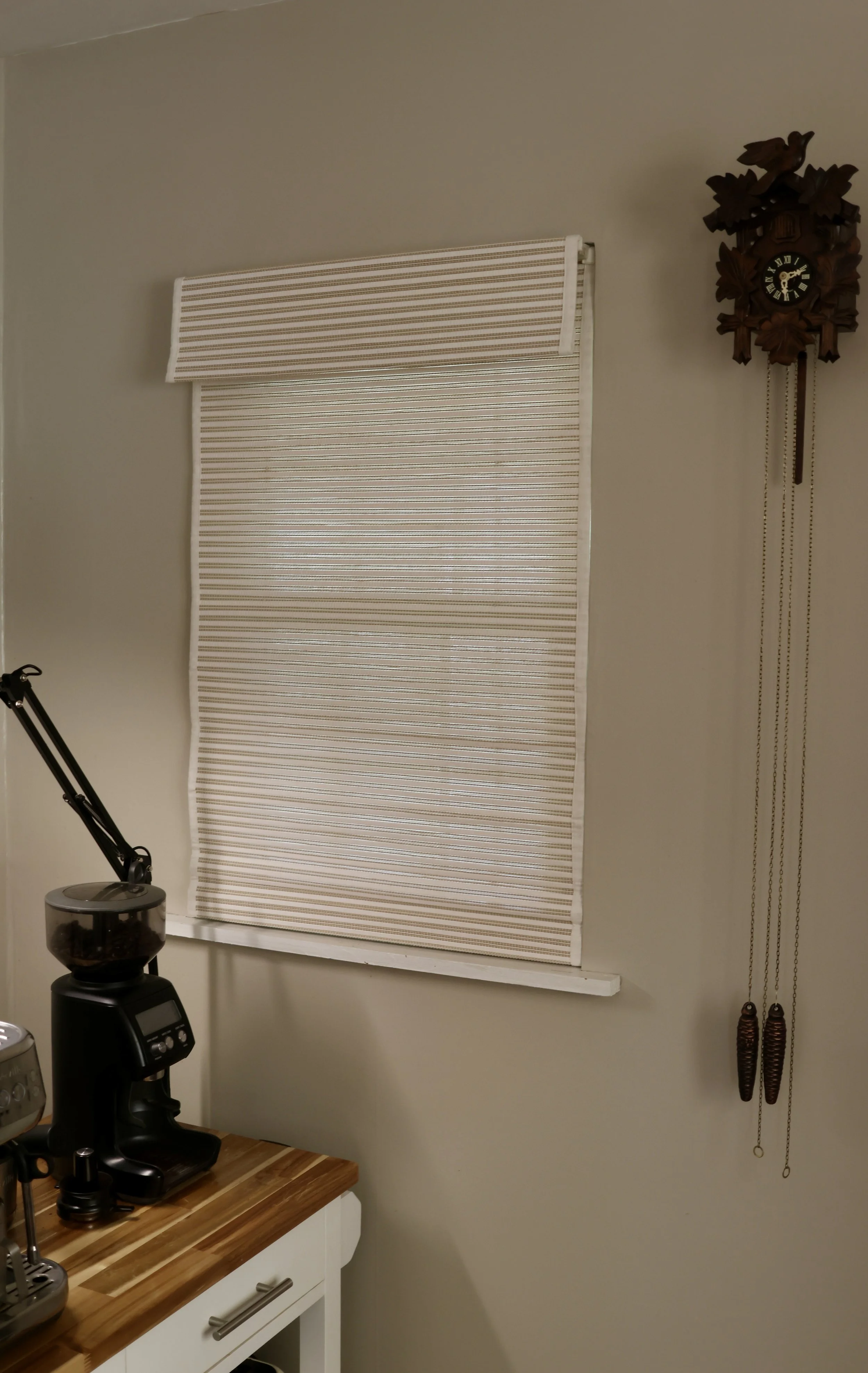 Interior of a room featuring a window with a beige and white striped roller shade, a vintage wooden cuckoo clock on the wall, a black kitchen appliance on a wooden counter, and a white cabinet with silver handles.