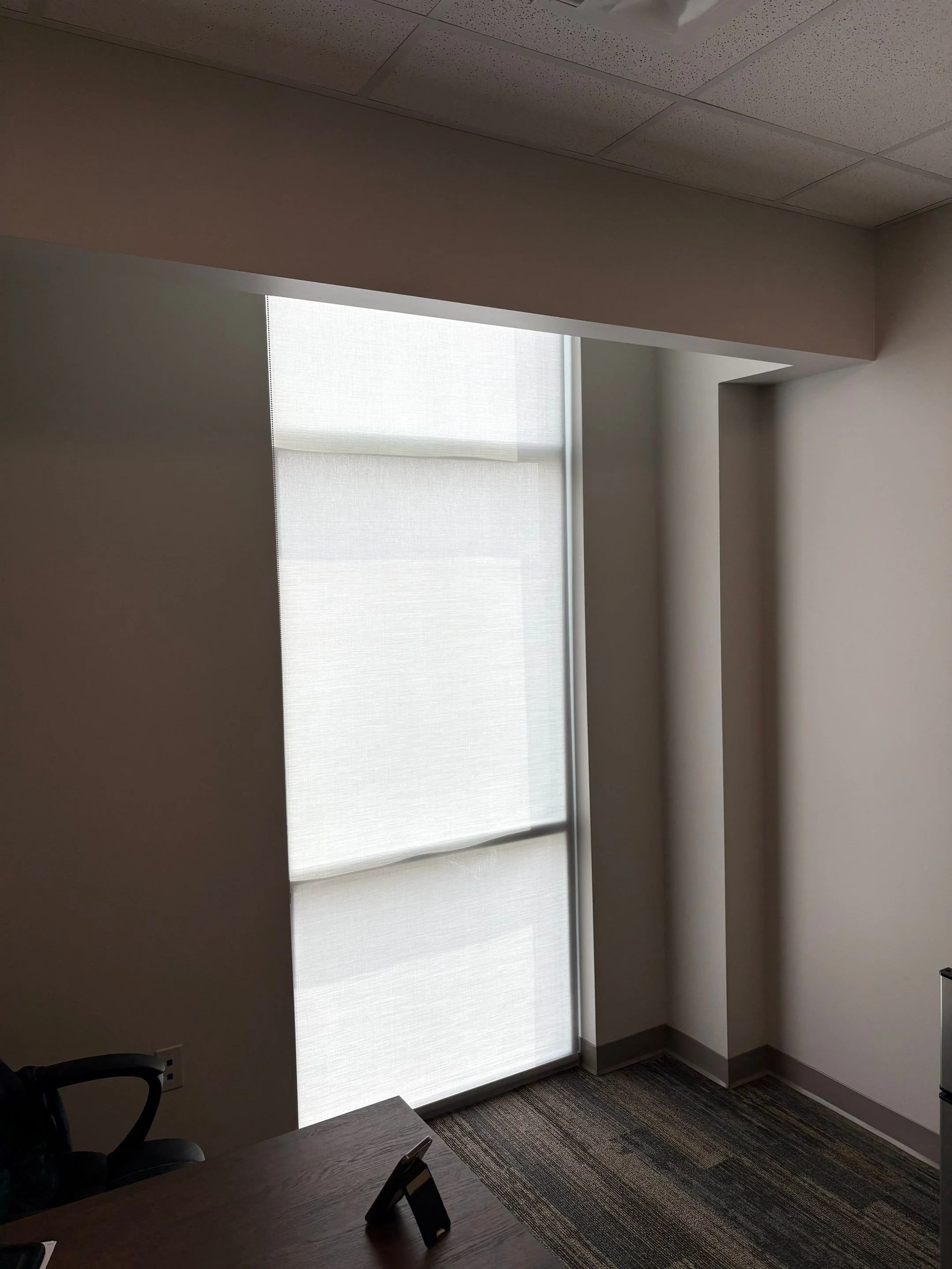 An office room with a large window covered by a white roller shade, part of a wooden desk with a phone on it, chairs, and a carpeted floor.
