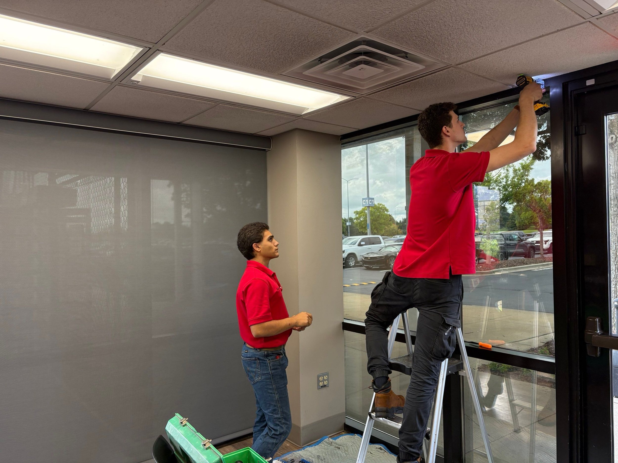 Two workers installing a ceiling vent in an office space. One worker is standing on a ladder, using a drill on the ceiling vent, while another is standing nearby watching. The room has large windows and fluorescent lights.