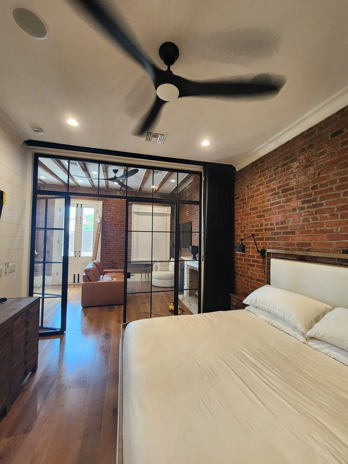 A bedroom with a white bed, brick wall, black wall lamp, wood nightstand, and a large square glass door leading to a sitting area with a sofa and a white armchair, and visible ceiling fan and skylight.