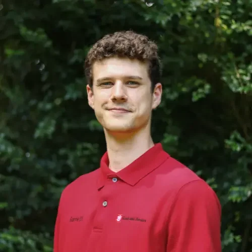 Young man with curly hair wearing a red polo shirt outdoors with green foliage in the background.
