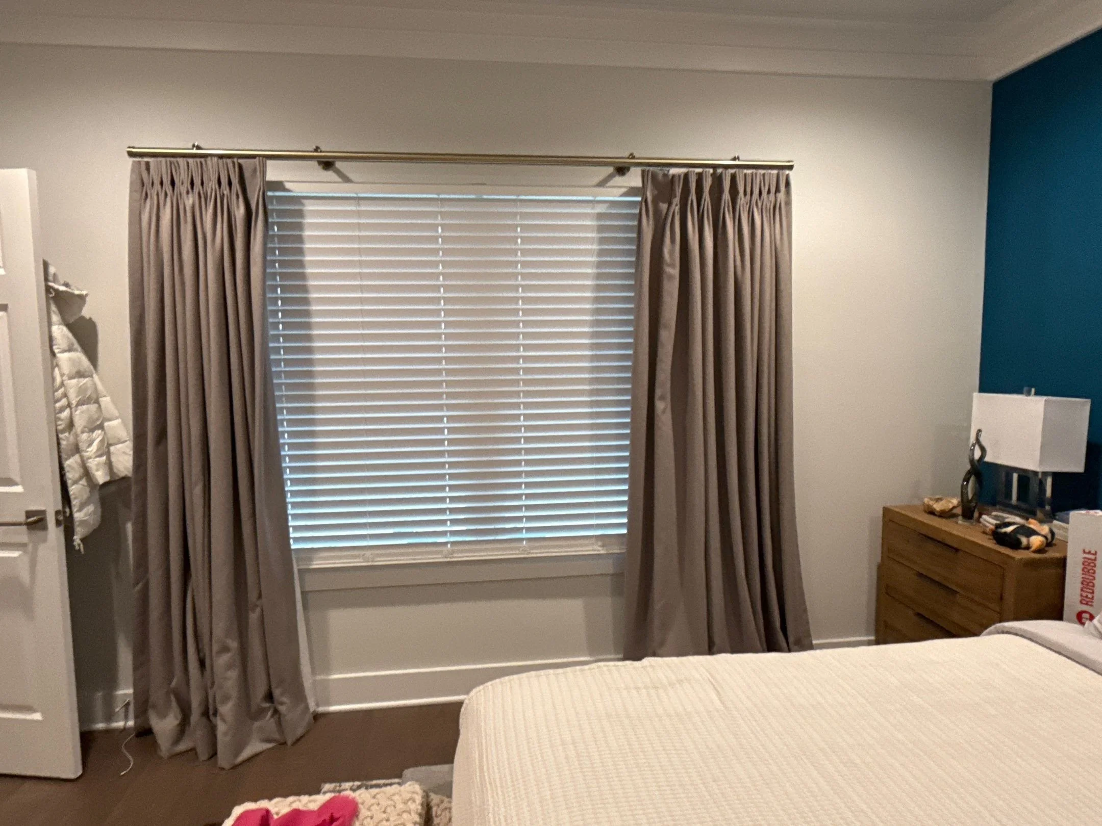 A bedroom with beige curtains and white blinds covering a window, a white quilted wall hanging on the left, a wooden nightstand with a few items including books and a decorative piece, and a bed with a white bedspread in the foreground. The walls are painted white and blue.