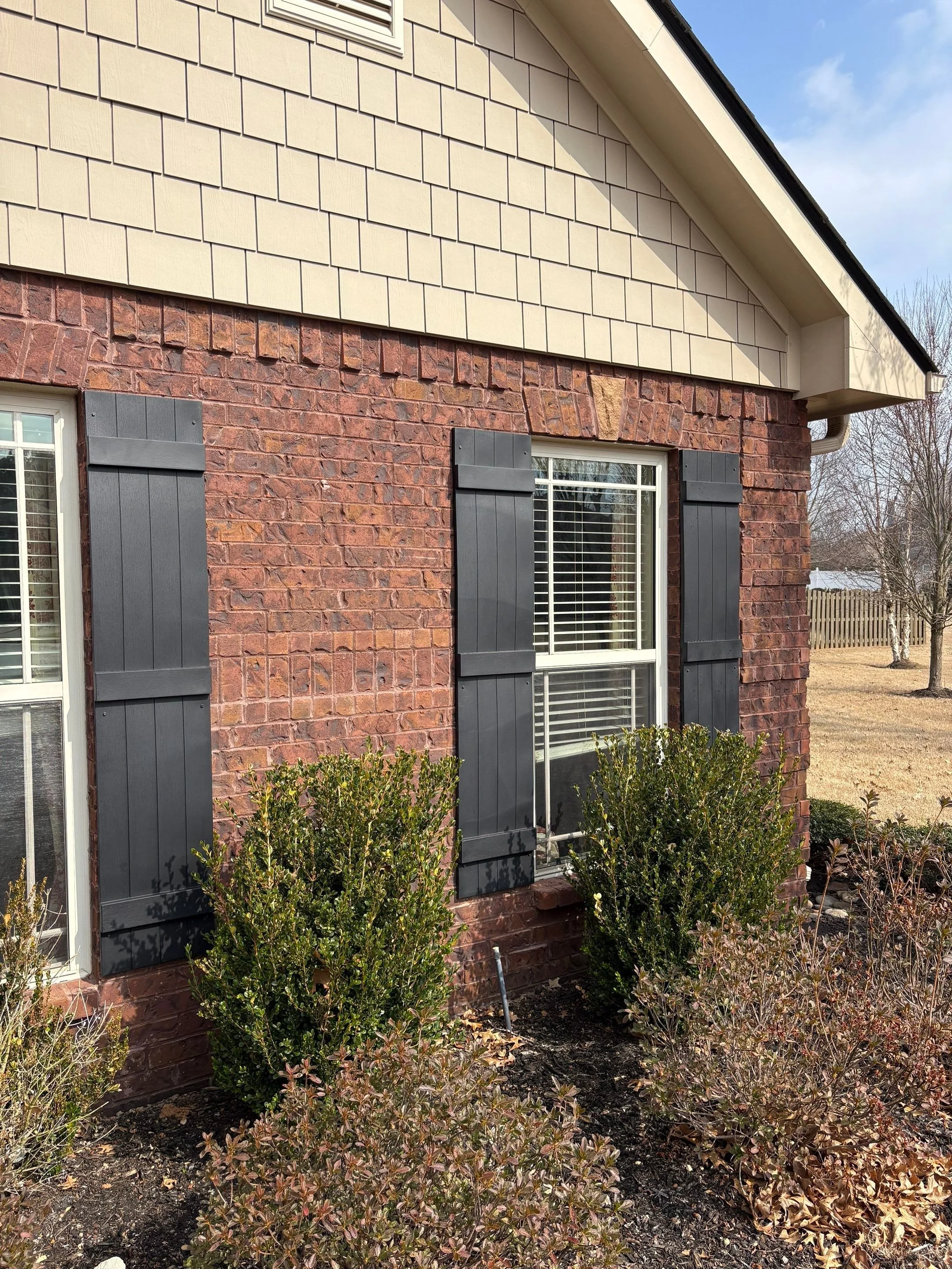 The image shows the exterior of a house with brick and beige siding. There are two windows with white frames and black shutters. Bushes are planted in front of the house, and the yard has some brown leaves and dirt. The sky is partly cloudy with some blue visible.