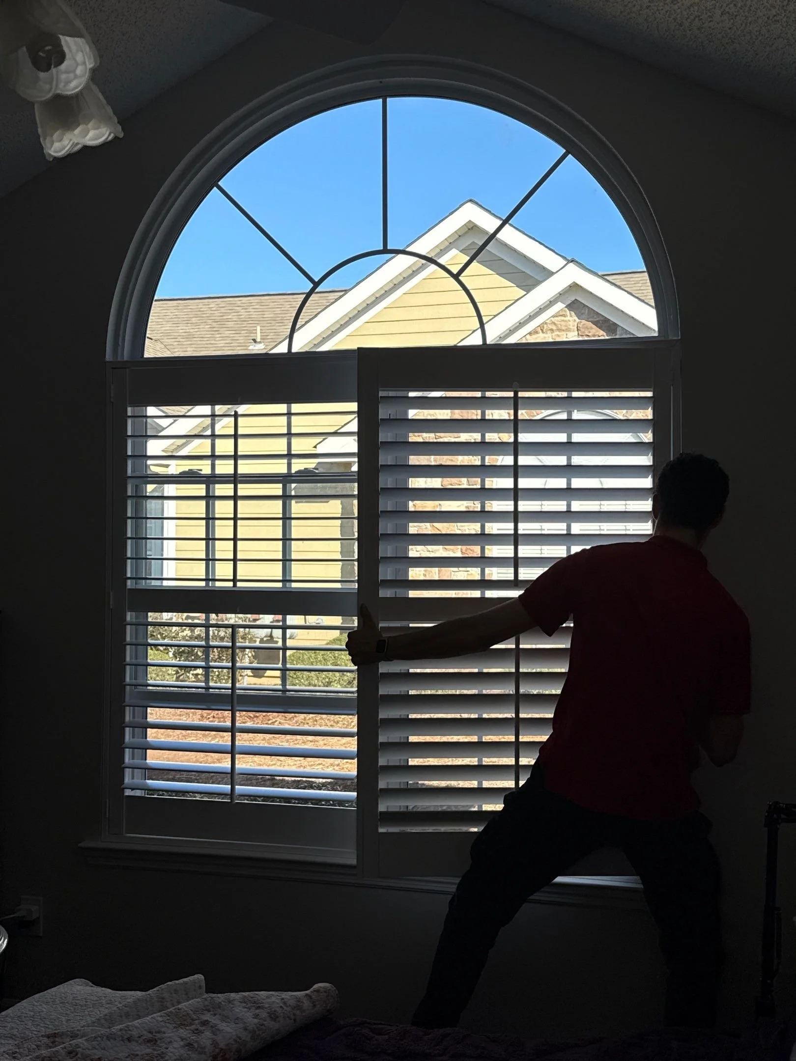 Person standing in front of a large window with shutters, with a blue sky and rooftops visible outside.