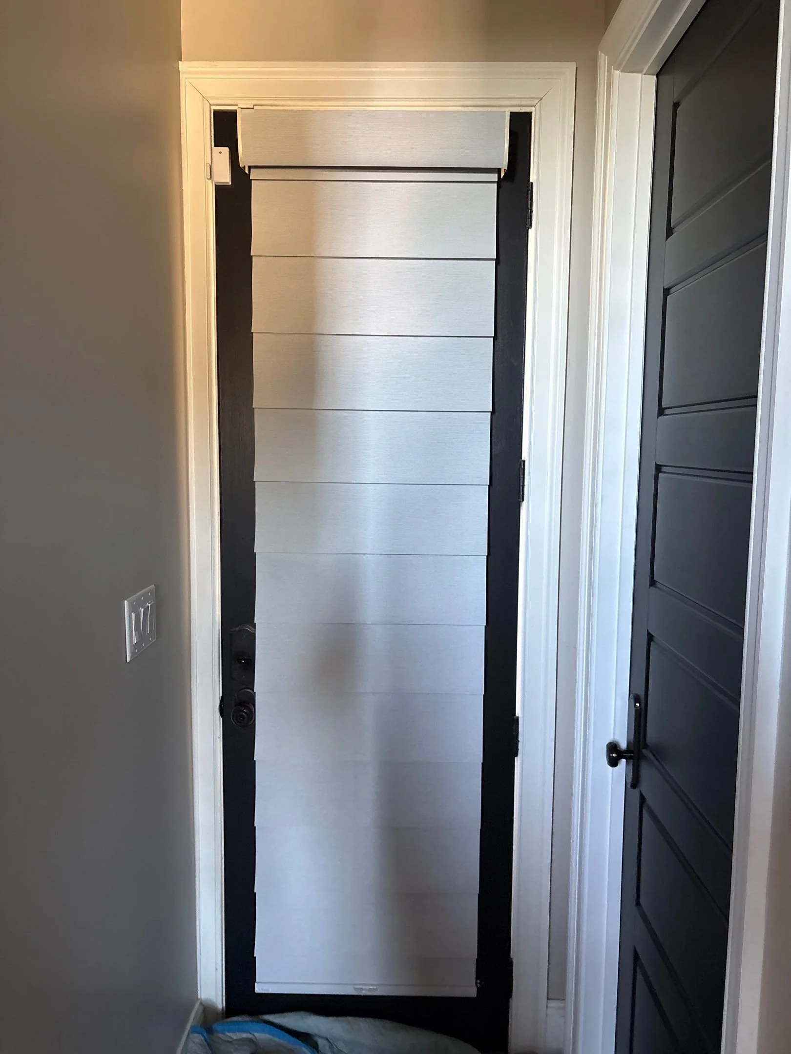 The image shows a black front door with a bottom and six horizontal white panels in the middle. To the right of the door, there is a dark gray wall with black paneling, and on the left wall, there is a white electrical outlet. The door frame is white, and the surrounding walls are painted light gray.