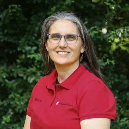 A woman with gray hair and glasses smiling outdoors with a leafy green background, wearing a red polo shirt.