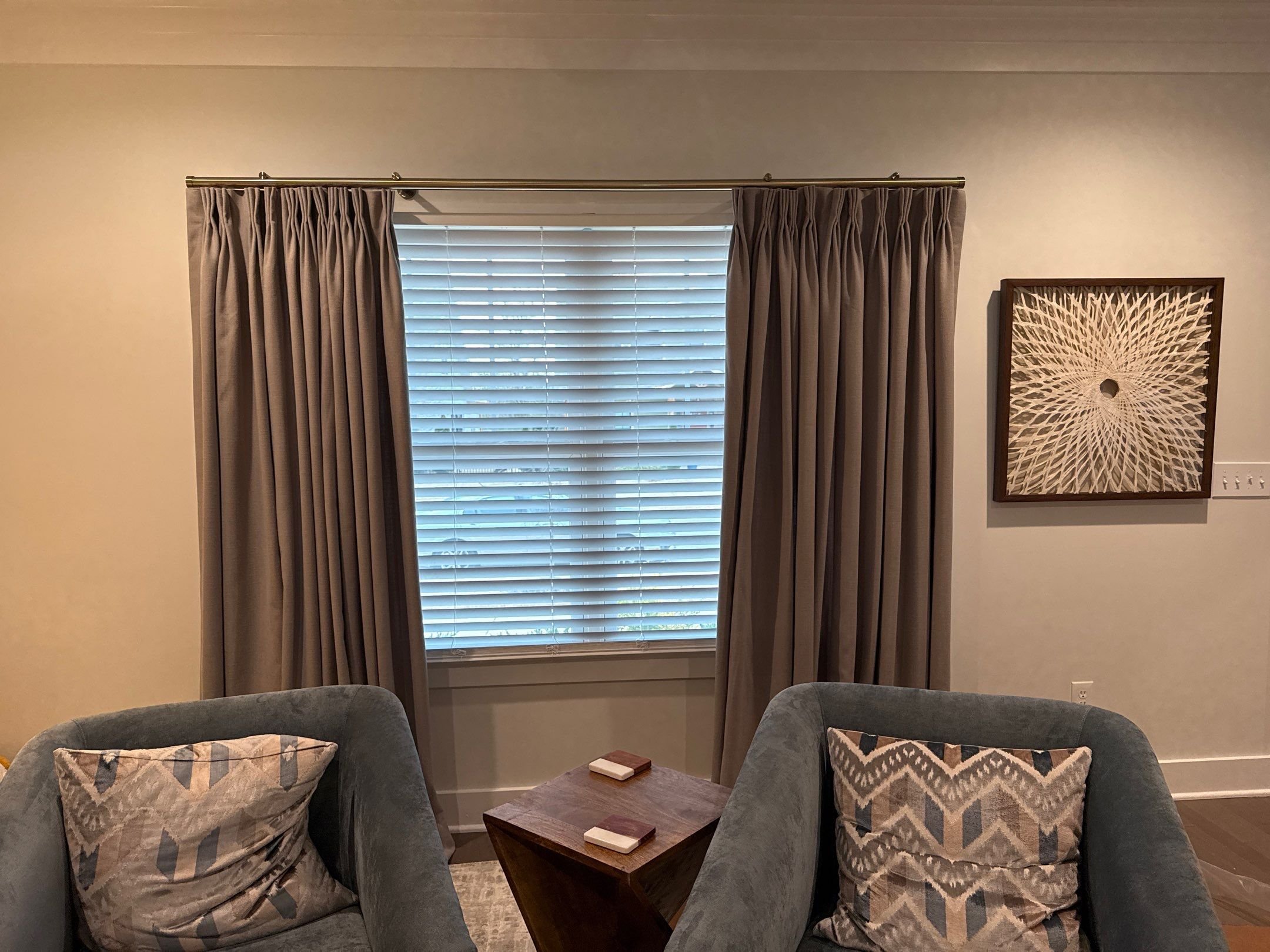 Living room with a window covered by beige curtains and white blinds, framed artwork on the right wall, and two gray armchairs with patterned pillows around a triangular wooden coffee table with coasters.