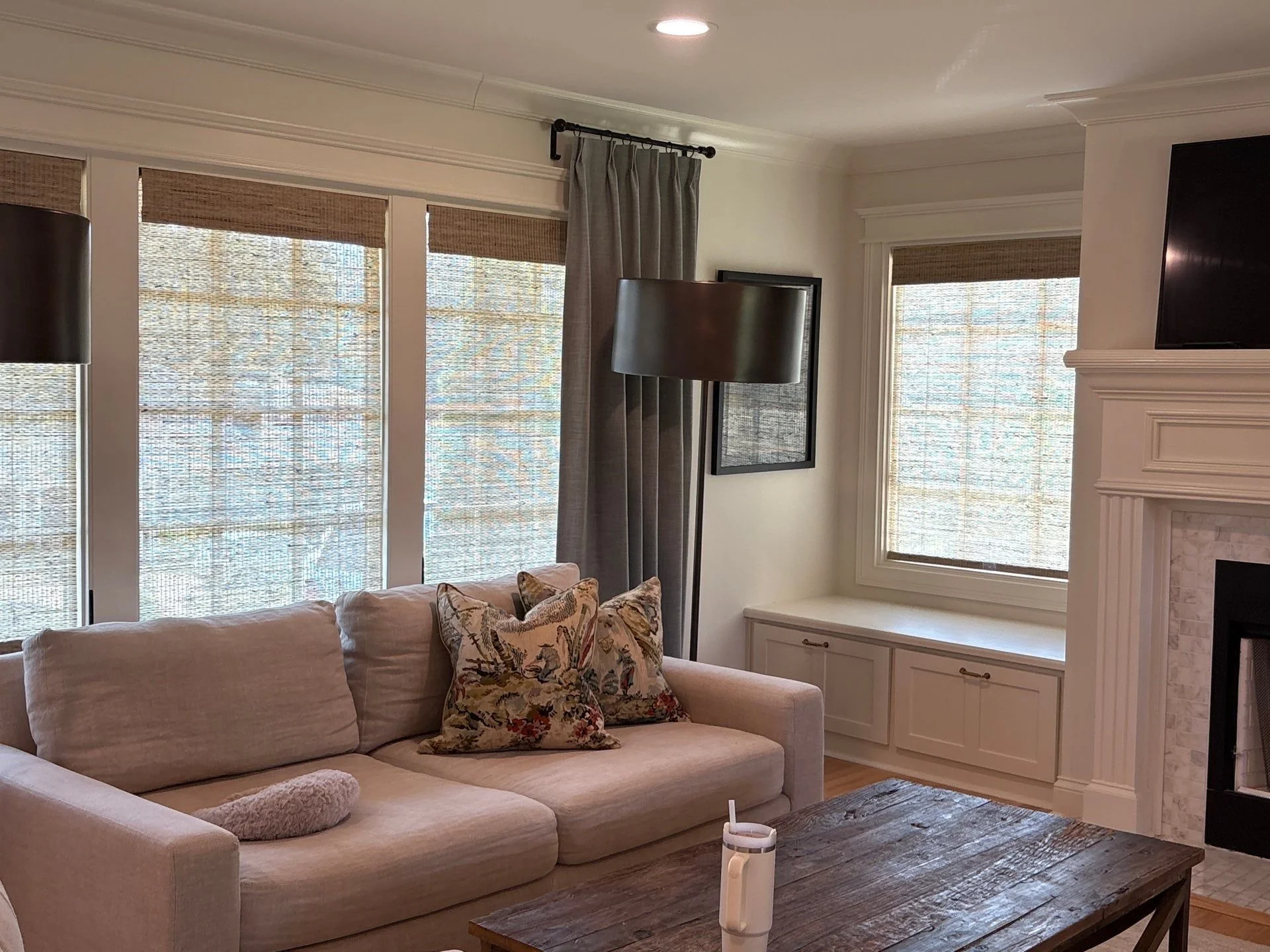 A living room with a beige sofa with floral and animal print pillows, a wooden coffee table, a white built-in cabinet below two windows with woven shades, a fireplace with a mantle, a television mounted above the fireplace, a floor lamp, and a corner window with beige curtains.