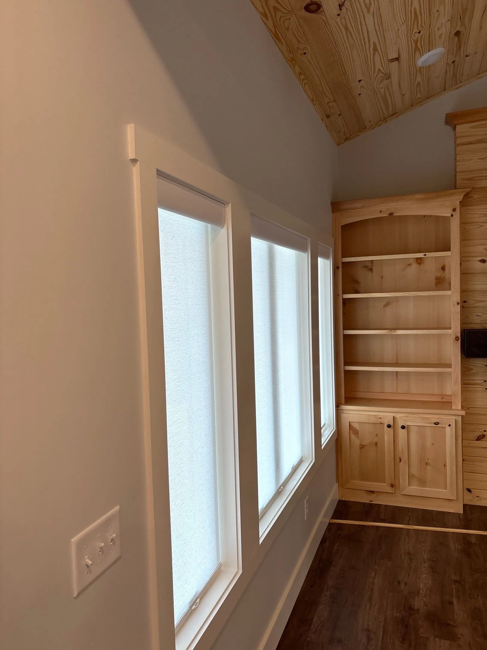 Interior of a room with three large windows with white frames and textured white blinds, white walls, wooden built-in shelves, and dark wood flooring.