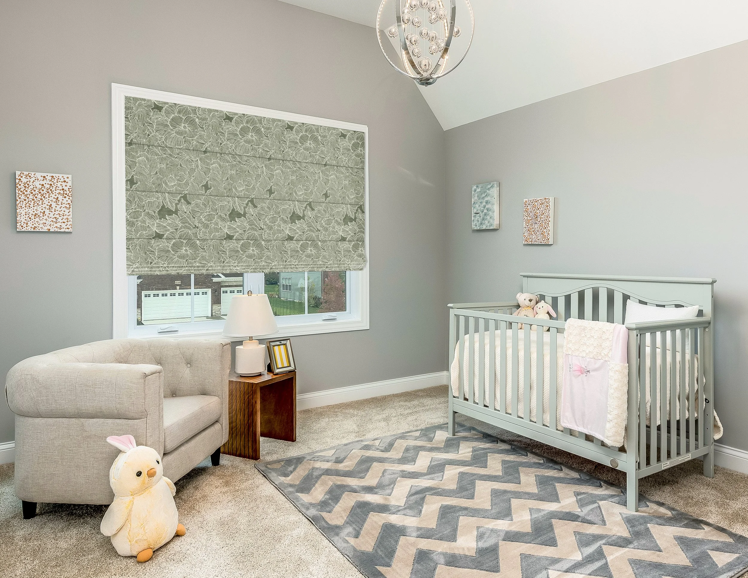 A nursery room with a gray wall, beige carpet, and a window with floral valance. Contains a beige armchair, a small wooden side table with a lamp and picture frame, a white crib with stuffed animals, a bunny plush toy on the floor, and a gray and blue chevron area rug. There is a round modern chandelier hanging from the ceiling and two small abstract wall art pieces.