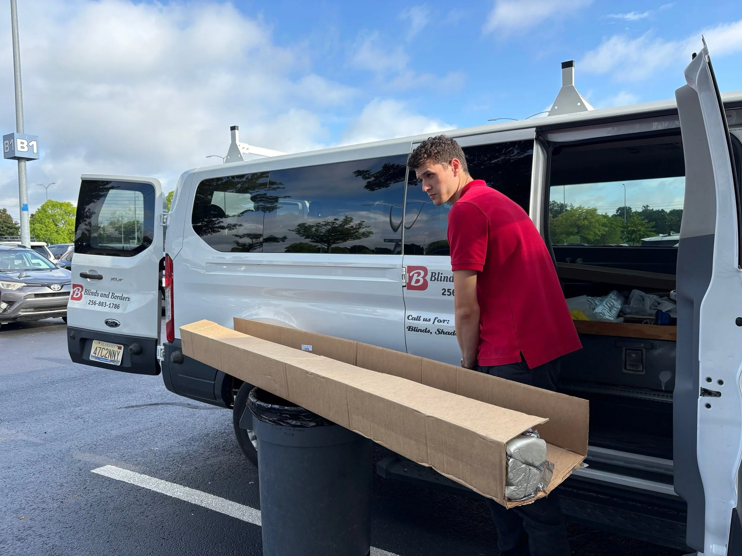 A young man in a red shirt loading a large cardboard box into the back of a white van at a parking lot.