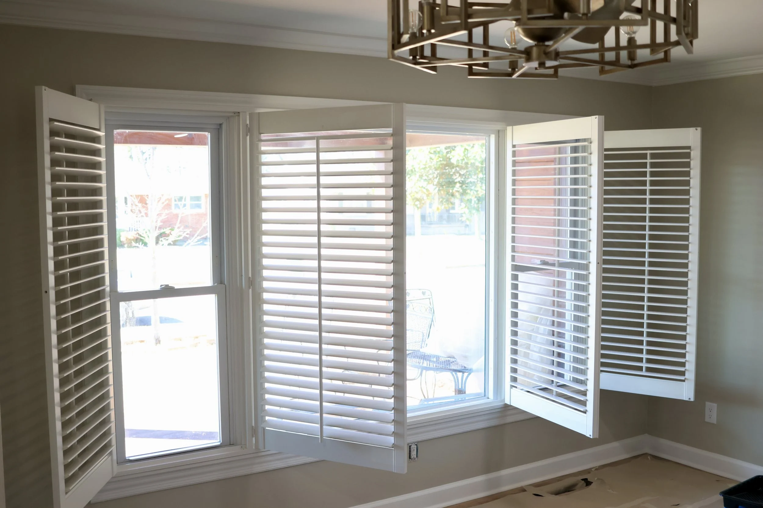 Close-up of white plantation shutters on large window, with sunlight coming through, revealing outdoor view of trees and a building.