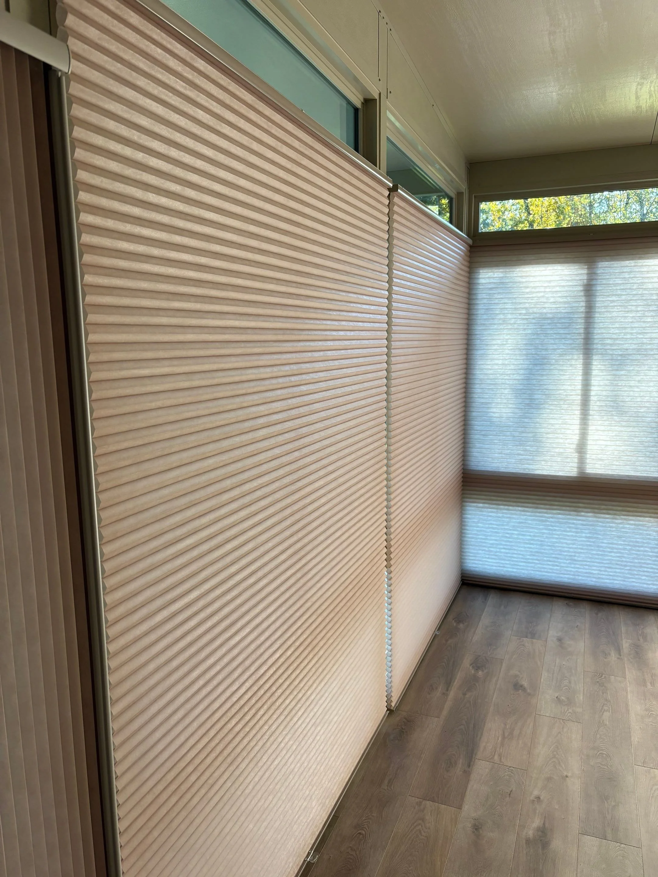 Close-up of beige pleated window blinds partially covering three multi-pane windows, with a view of trees outside, and a floor with wooden pattern.