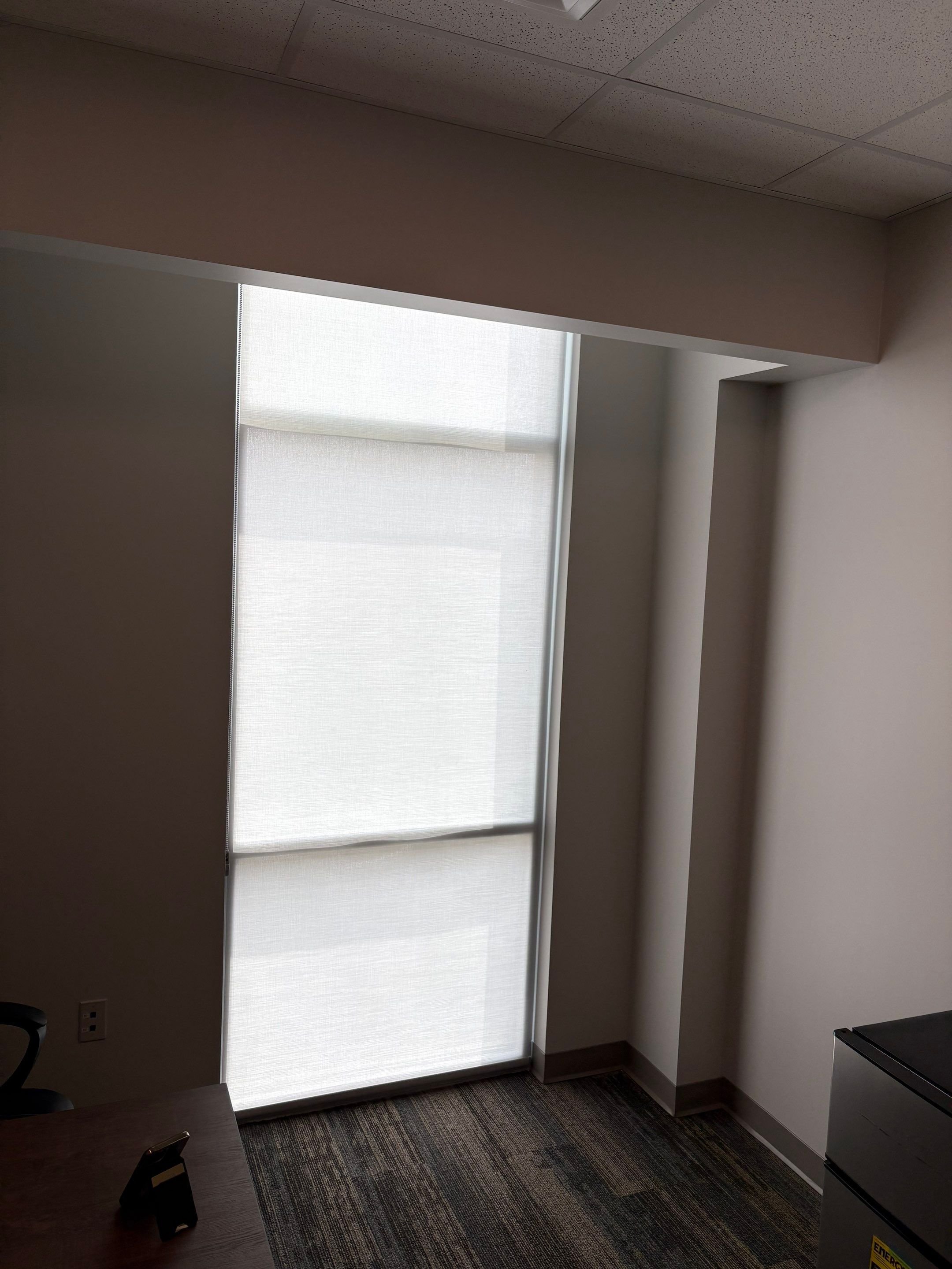 Interior view of a room with a window covered by a white blind, a ceiling with acoustic tiles, a wall with an electrical outlet, and part of a desk or table in the foreground.
