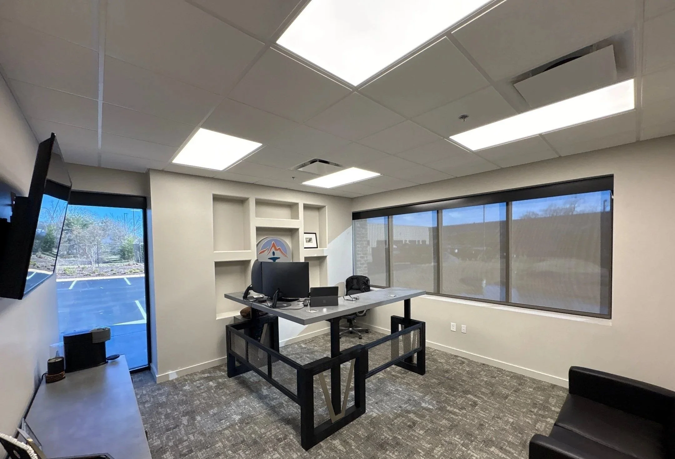 An empty office with large windows and a ceiling with bright fluorescent lights. There is a desk with a chair, a computer monitor, and some office supplies. The office has a gray carpet and beige walls, with a parking lot and trees visible outside.