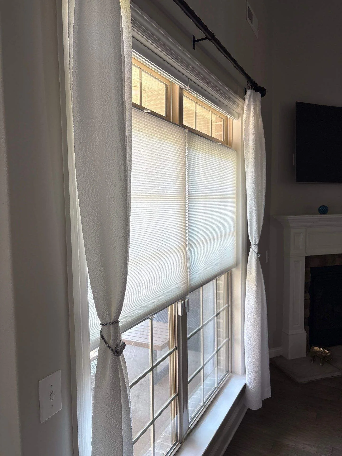 Living room window with white textured curtains pulled to the sides and white blinds partially closed, sunlight shining through.