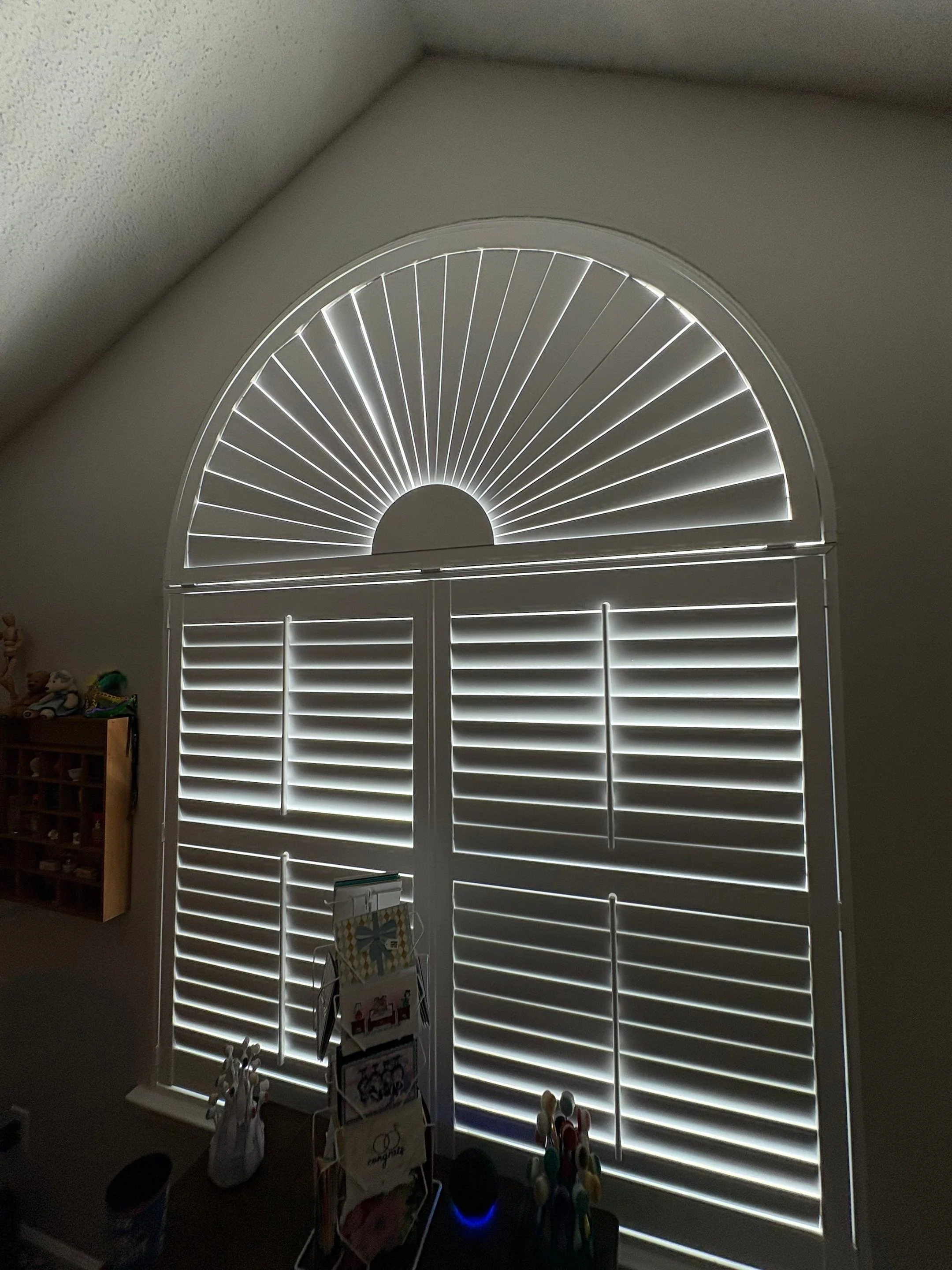 Sunlight streaming through a decorative semi-circular window with white shutters in a room. The room has some toys and objects, including a rack with blank cards and some spools of thread or ribbon on a table.