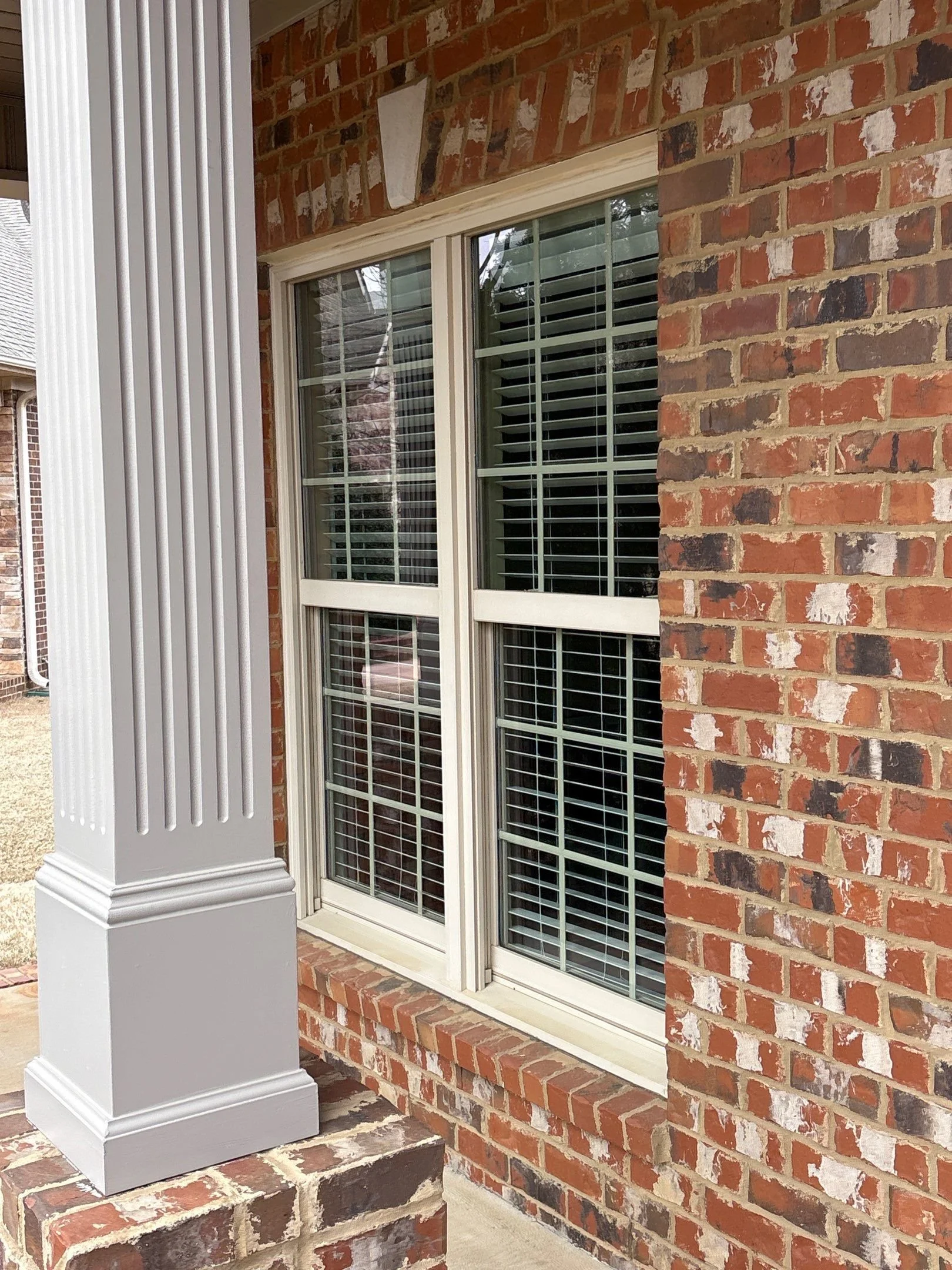 Close-up of a brick house exterior with a window with white trim, white lattice blinds, and white column with decorative base.