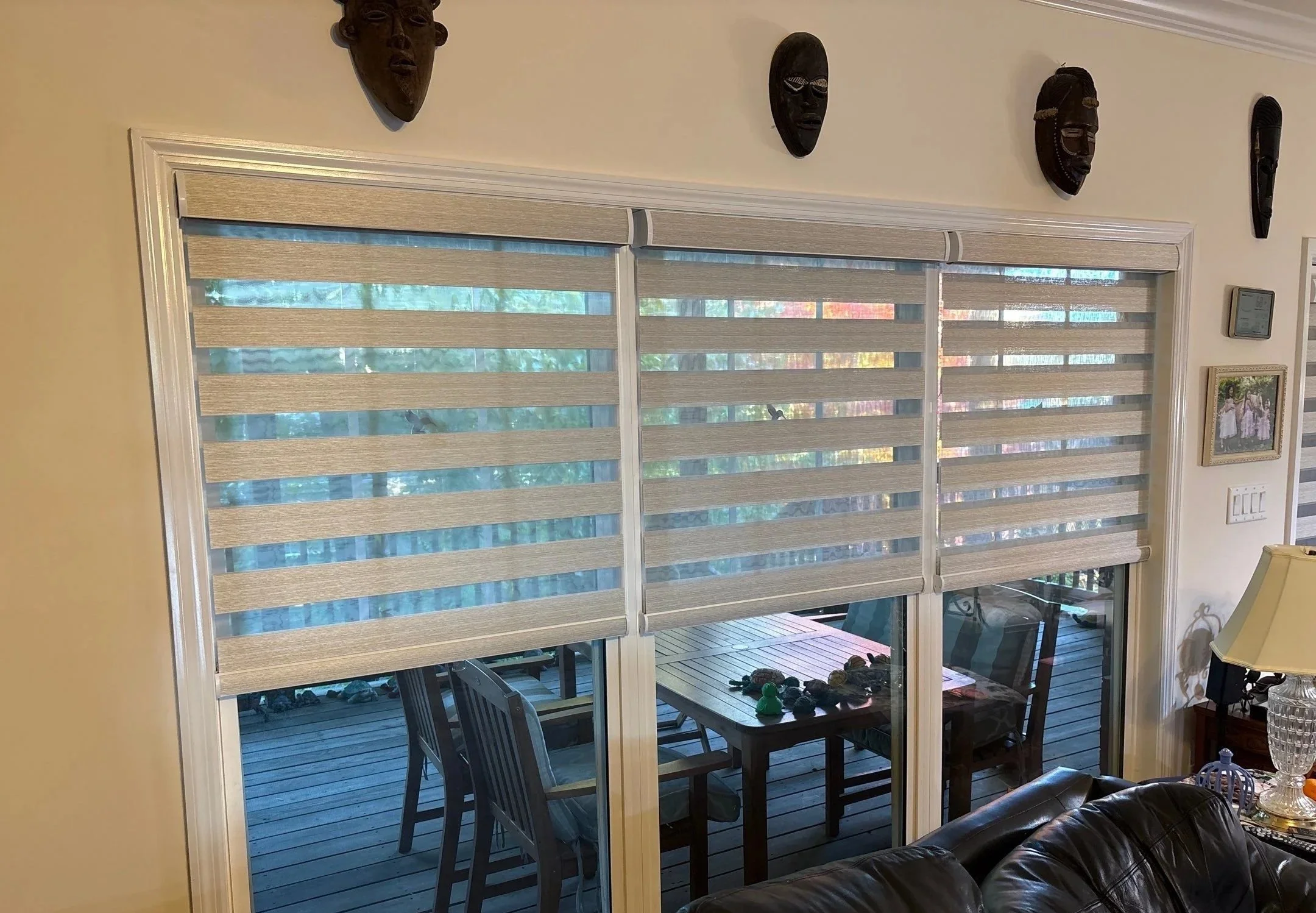 View of a covered outdoor patio area through sliding glass doors with beige blinds. The patio has a wooden deck, black chairs, and a table with a green toy on it. The interior room has beige walls, tribal masks on the wall, a thermostat, a framed picture, a table lamp, and a leather sofa.