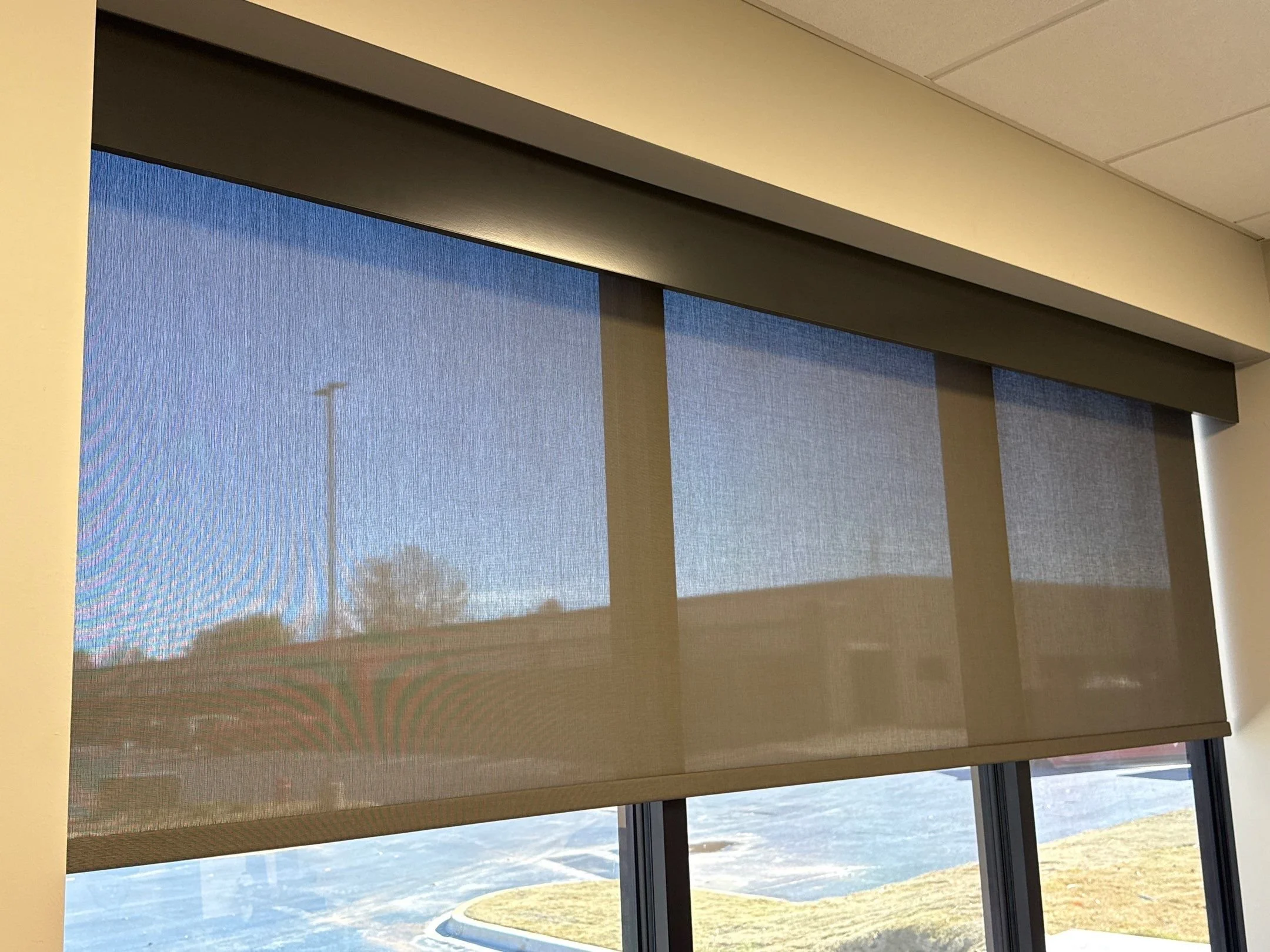 Indoor view of closed window blinds covering large windows with a view of a parking lot and grass outside.