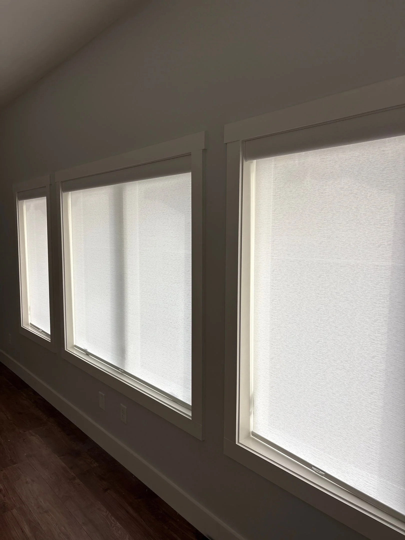 Three windows with closed white blinds in a room with dark wooden floor.