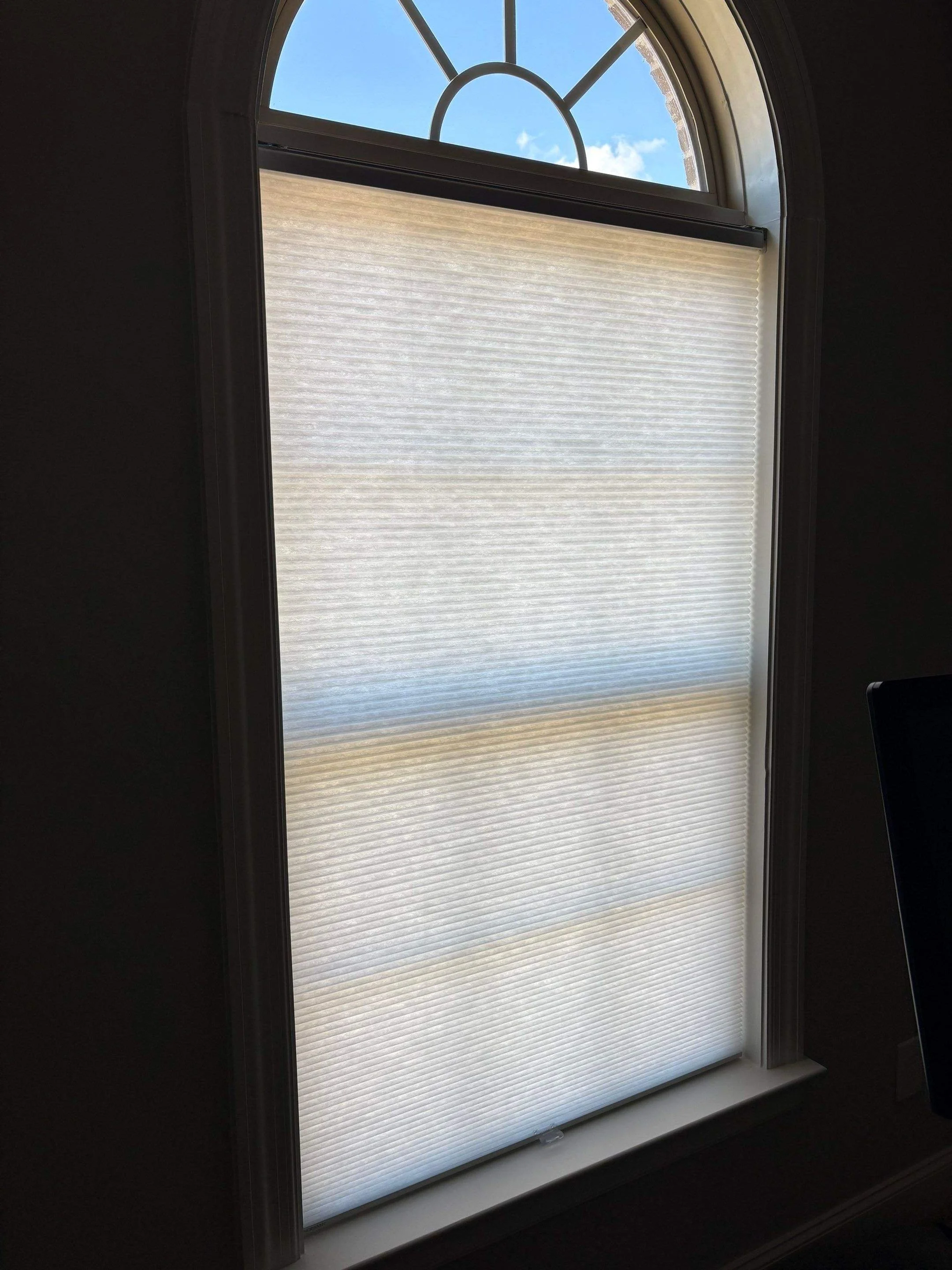 A partially open window with a semi-circular top, showing blue sky with a few clouds outside. There is a white pleated shade covering the lower part of the window.