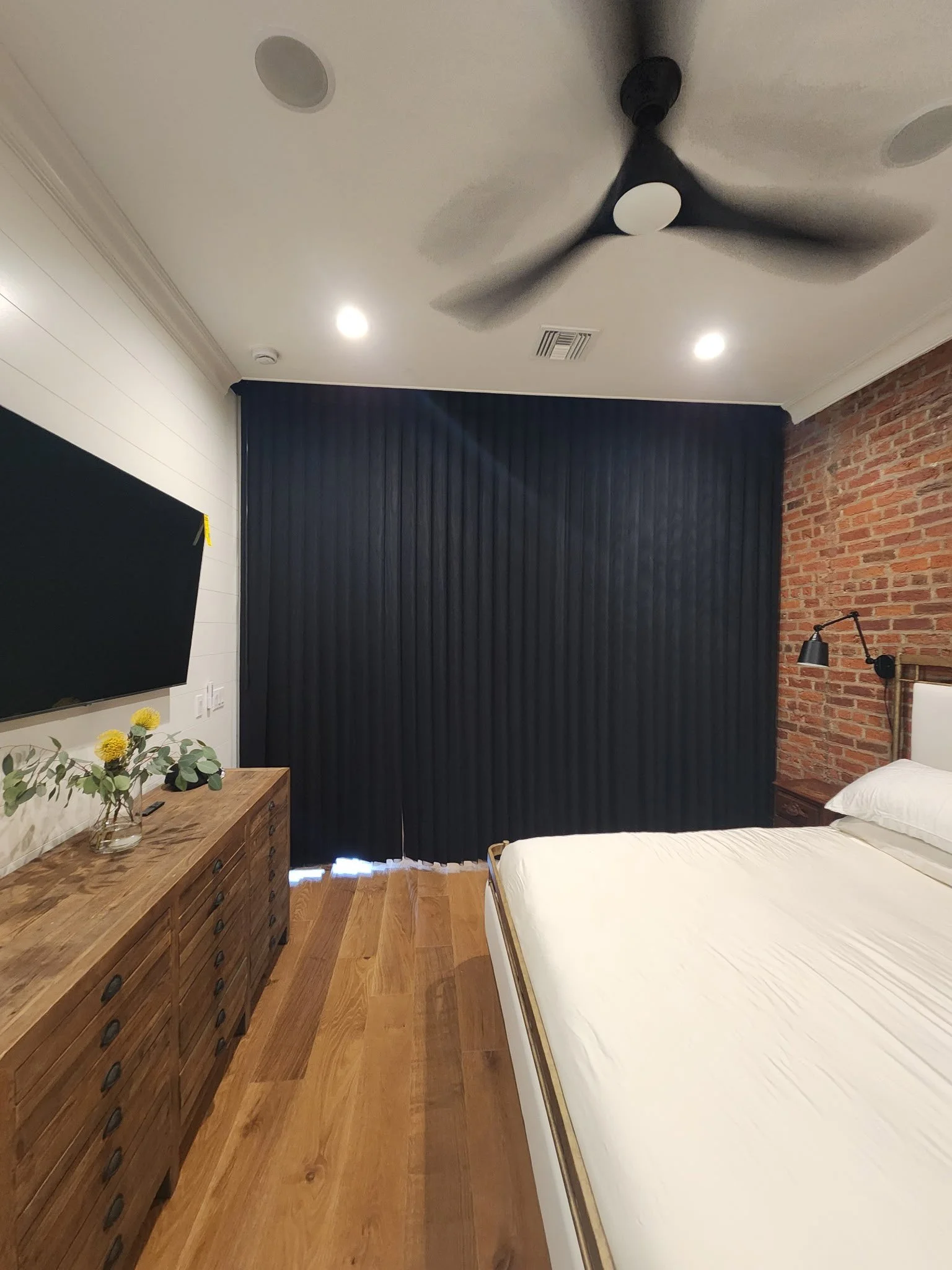 A bedroom with a wooden dresser, a bed with white bedding, a black curtain covering a window, a brick wall, a wall-mounted lamp, and a ceiling fan with three blades.