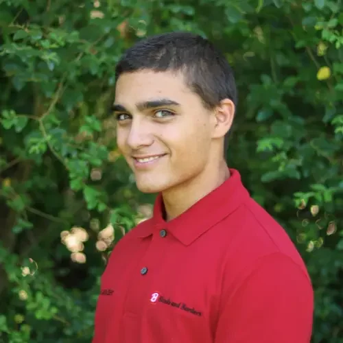 Young man with short dark hair, wearing a red polo shirt, smiling outdoors with green foliage in the background.