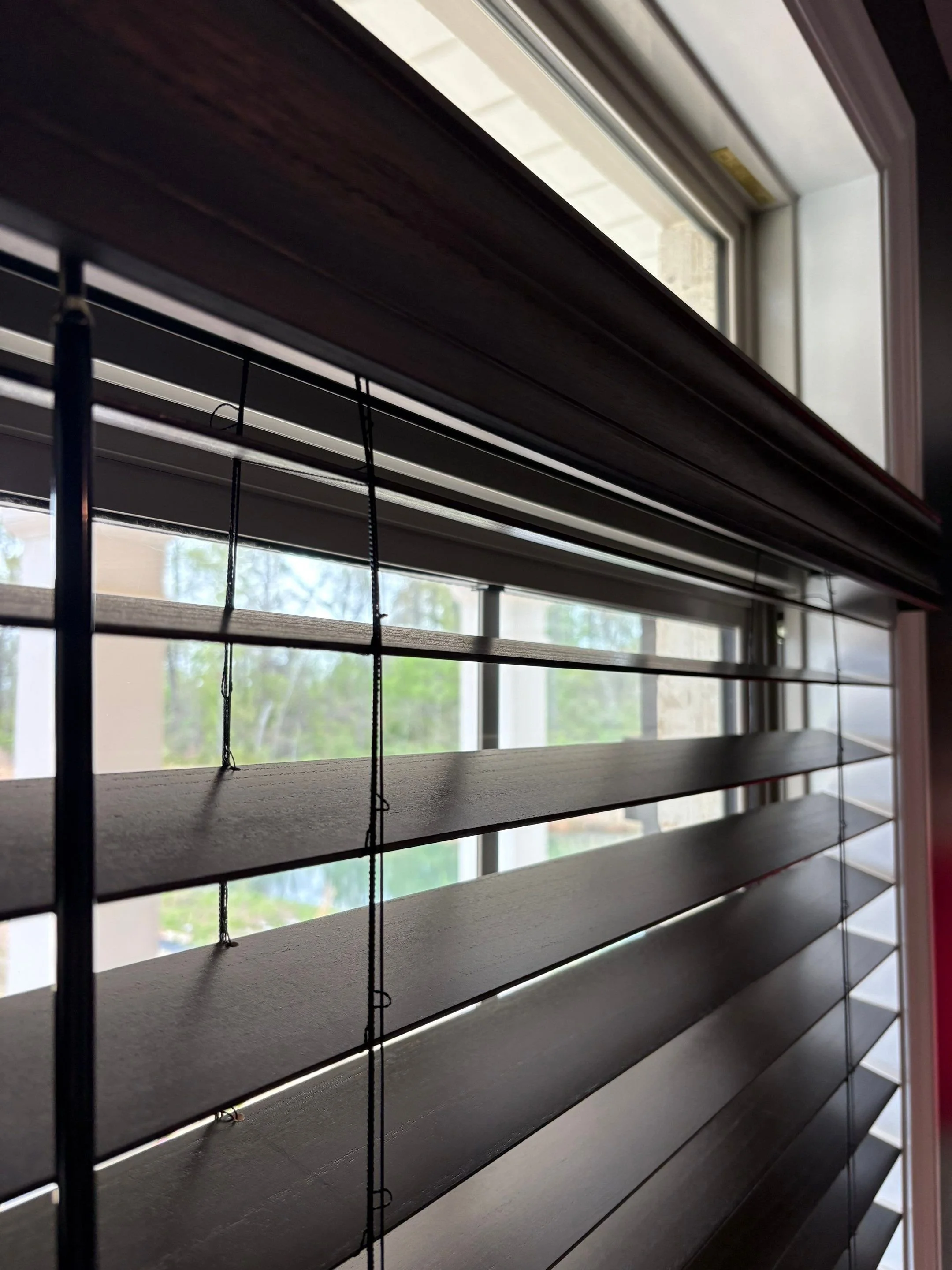 Close-up view of window blinds with horizontal slats, partially opened, with a window and outdoor scene visible through the gaps.