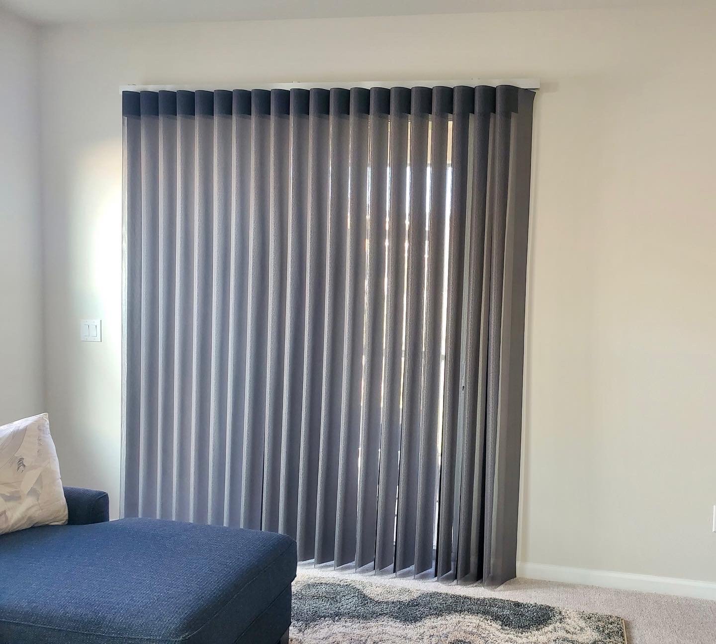 A living room with closed vertical blinds covering a window. The wall is painted white, and there's a dark blue sofa with a pillow and a patterned area rug on the light-colored carpeted floor.