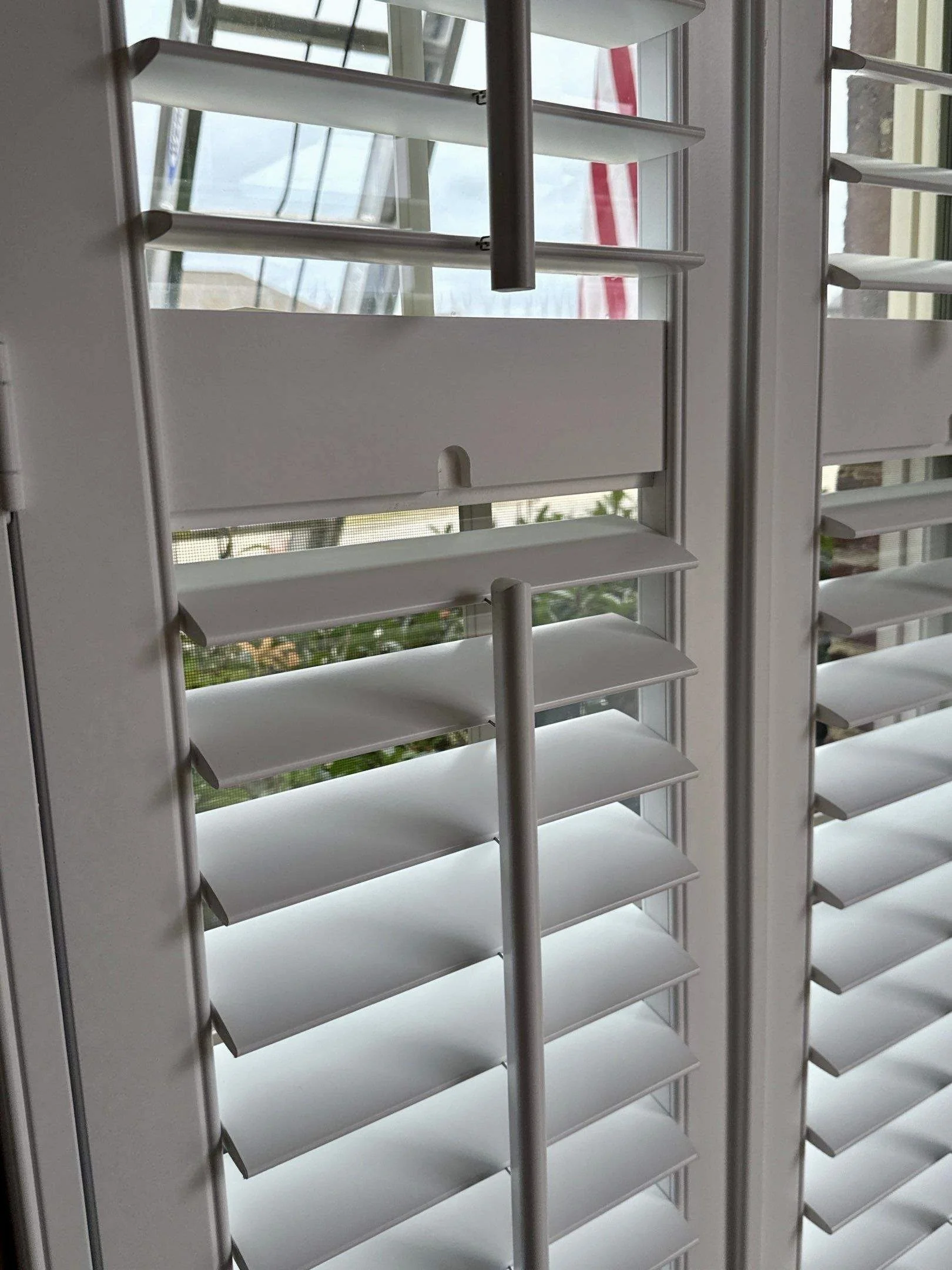 White window blinds partially open, revealing a window and an outdoor scene with a red and white striped object and some greenery.