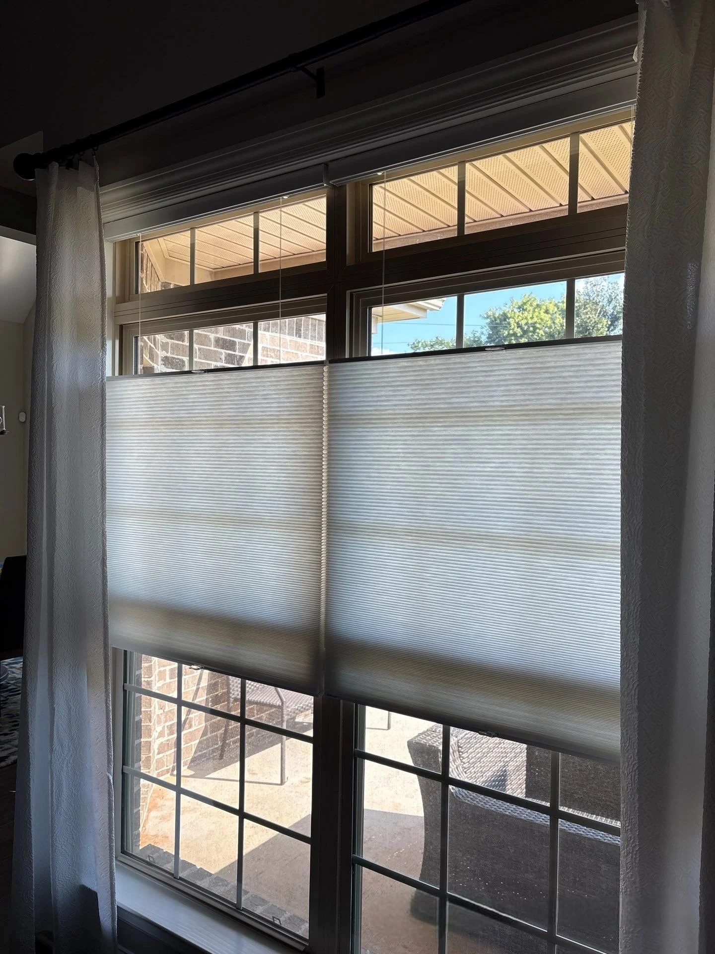 A window with a white cellular shade partially pulled down, outside view of a brick wall and greenery, with a curtain on the left side.