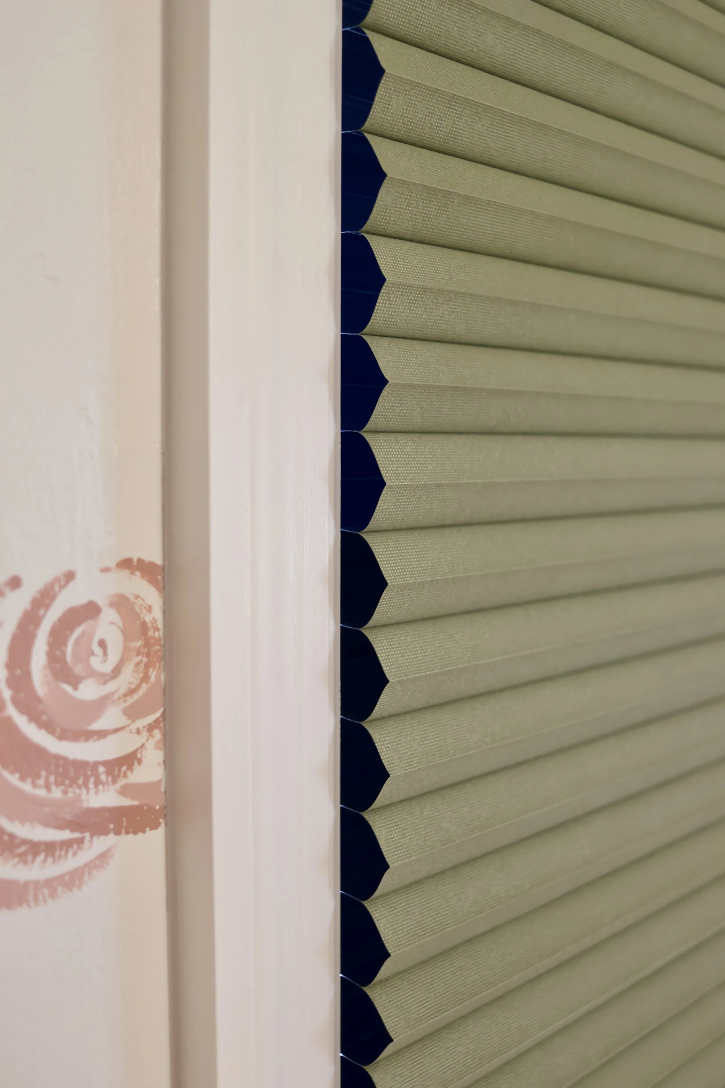 A window with closed beige blinds with black tips, and a white windowsill with a pink floral design on the left side.