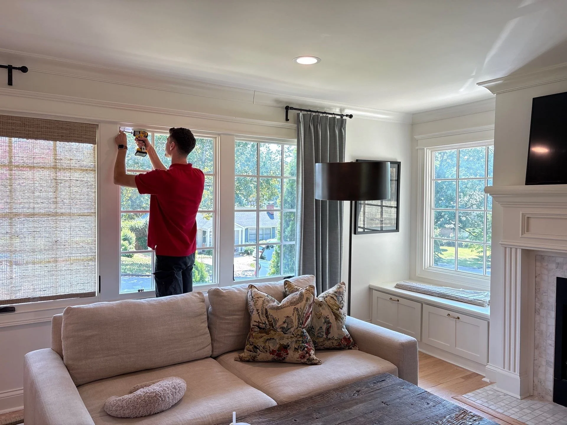 A man in a red shirt is using a cordless drill to install a window blind on a large window in a bright living room. The room features white walls, crown molding, a beige sofa with patterned pillows, a floor lamp, and a flat-screen TV mounted on the wall. Outside the window, trees and neighborhood houses are visible.