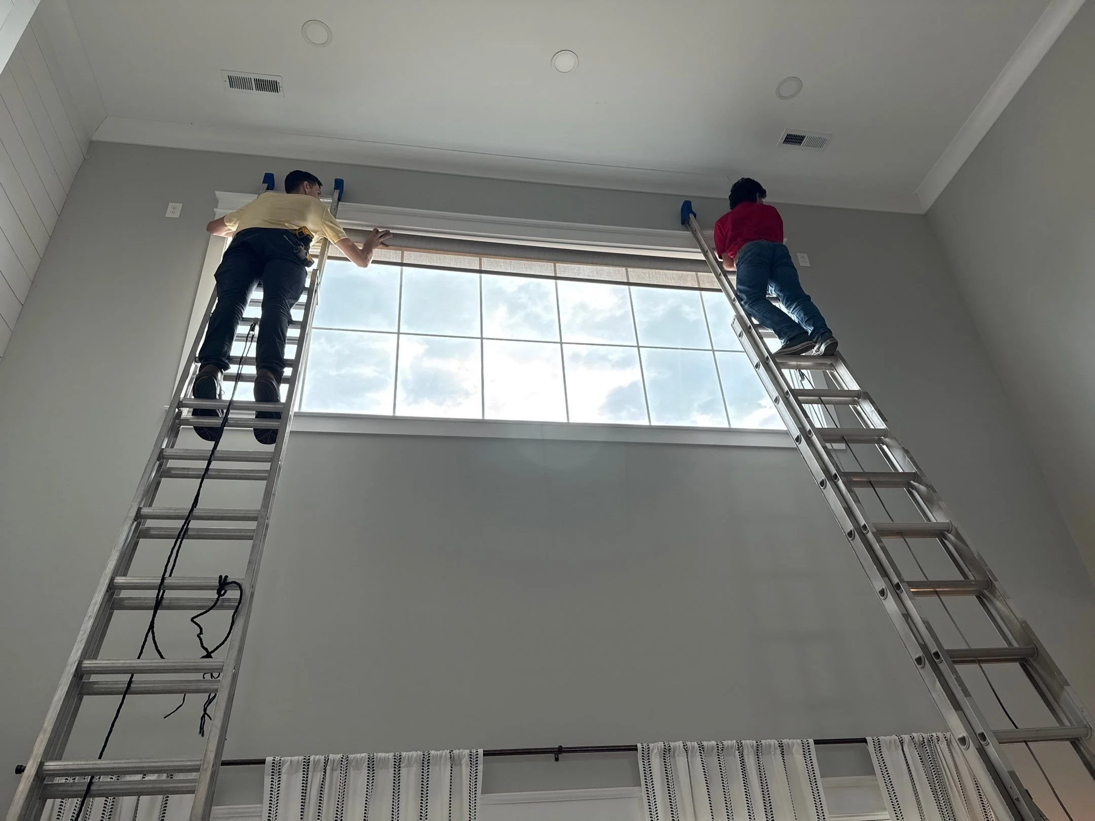 Two workers are on ladders installing or adjusting a large window in a room with white walls and ceiling. The window shows a view of a cloudy sky.