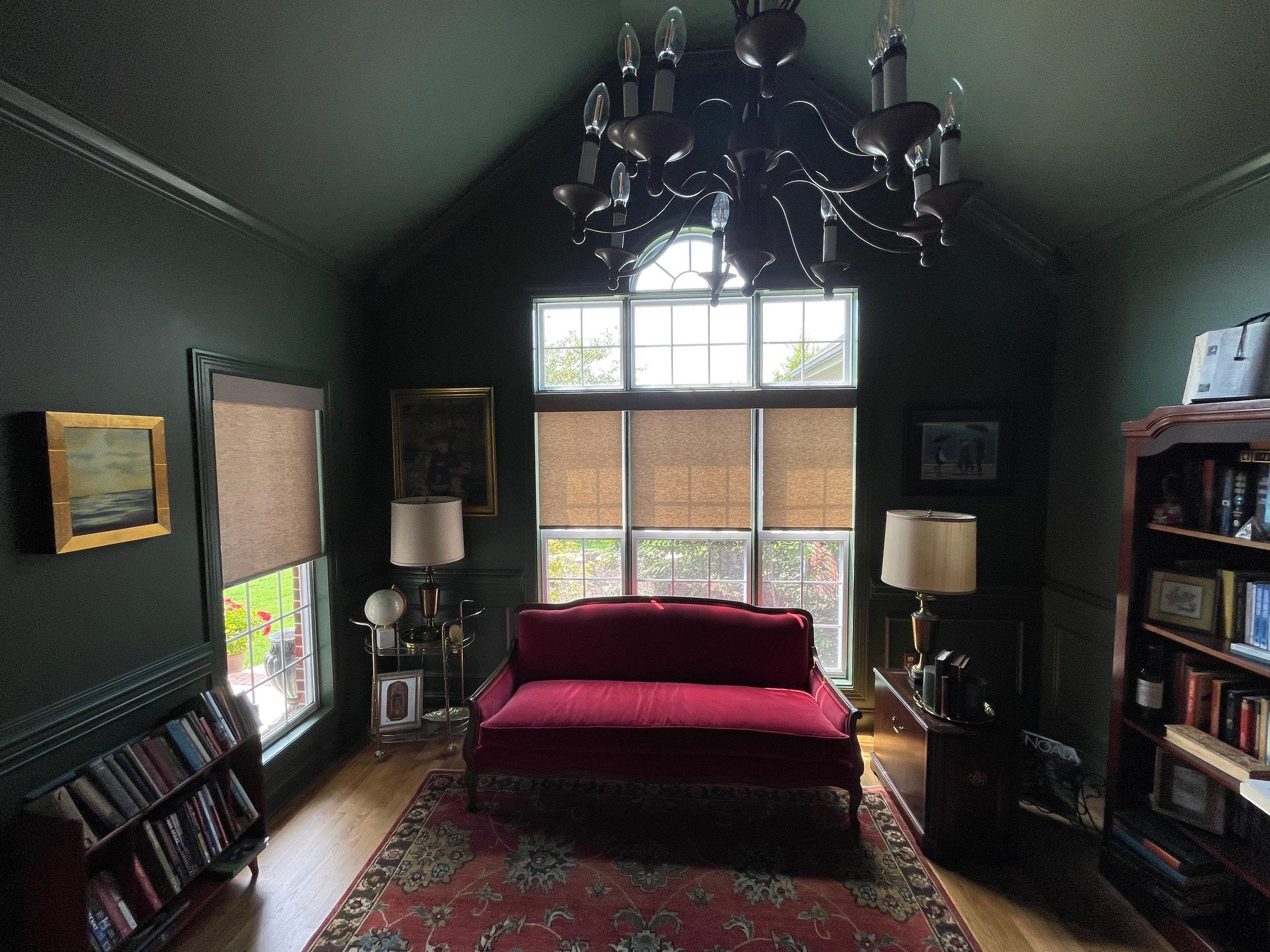 A dark green living room with tall windows, a red velvet sofa, a patterned area rug, and a black chandelier hanging from the ceiling.