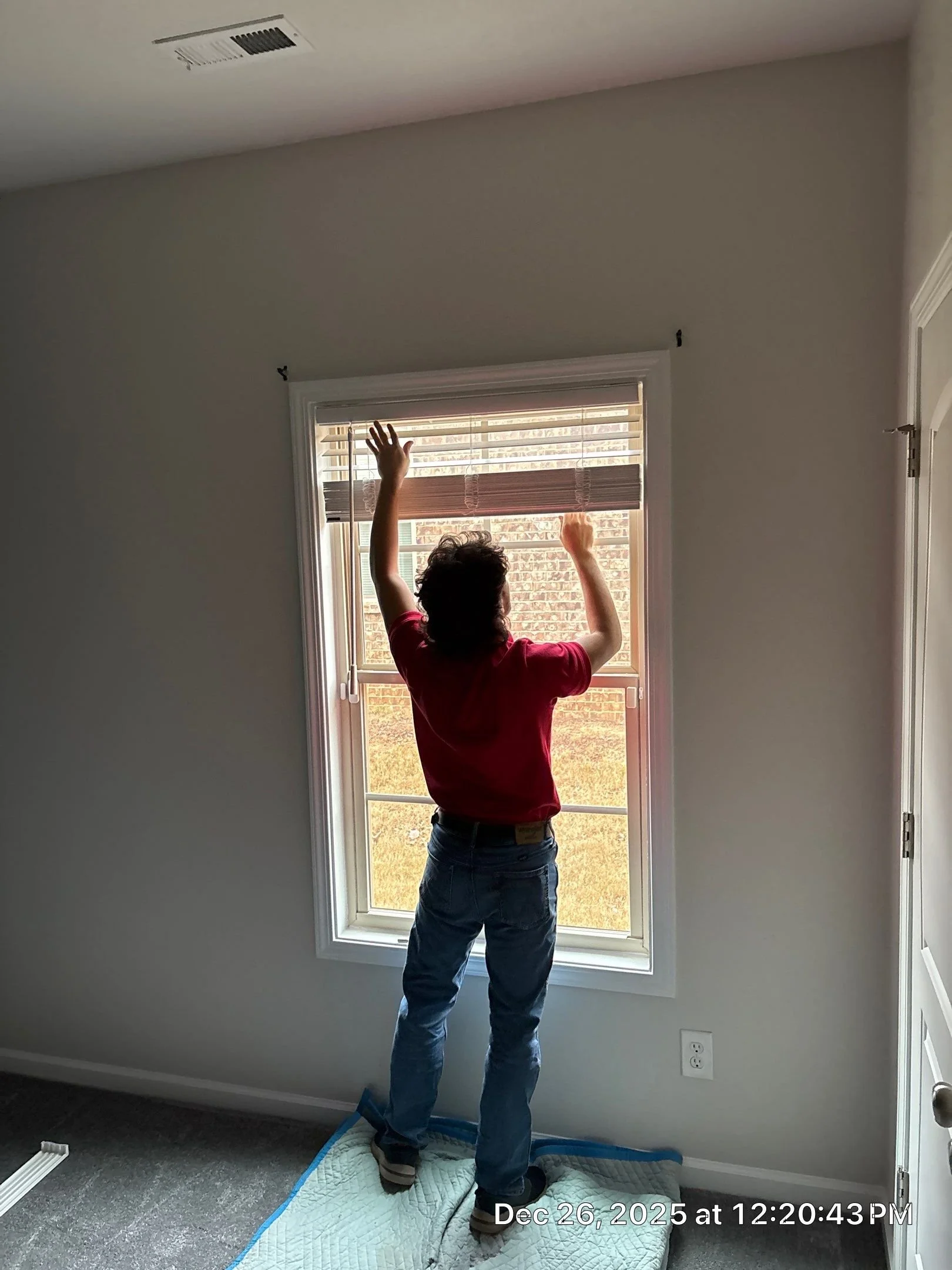 A person standing on a bed while adjusting window blinds in a room with gray walls, a large window, and a view of brick exterior. The person is wearing a red shirt and blue jeans.