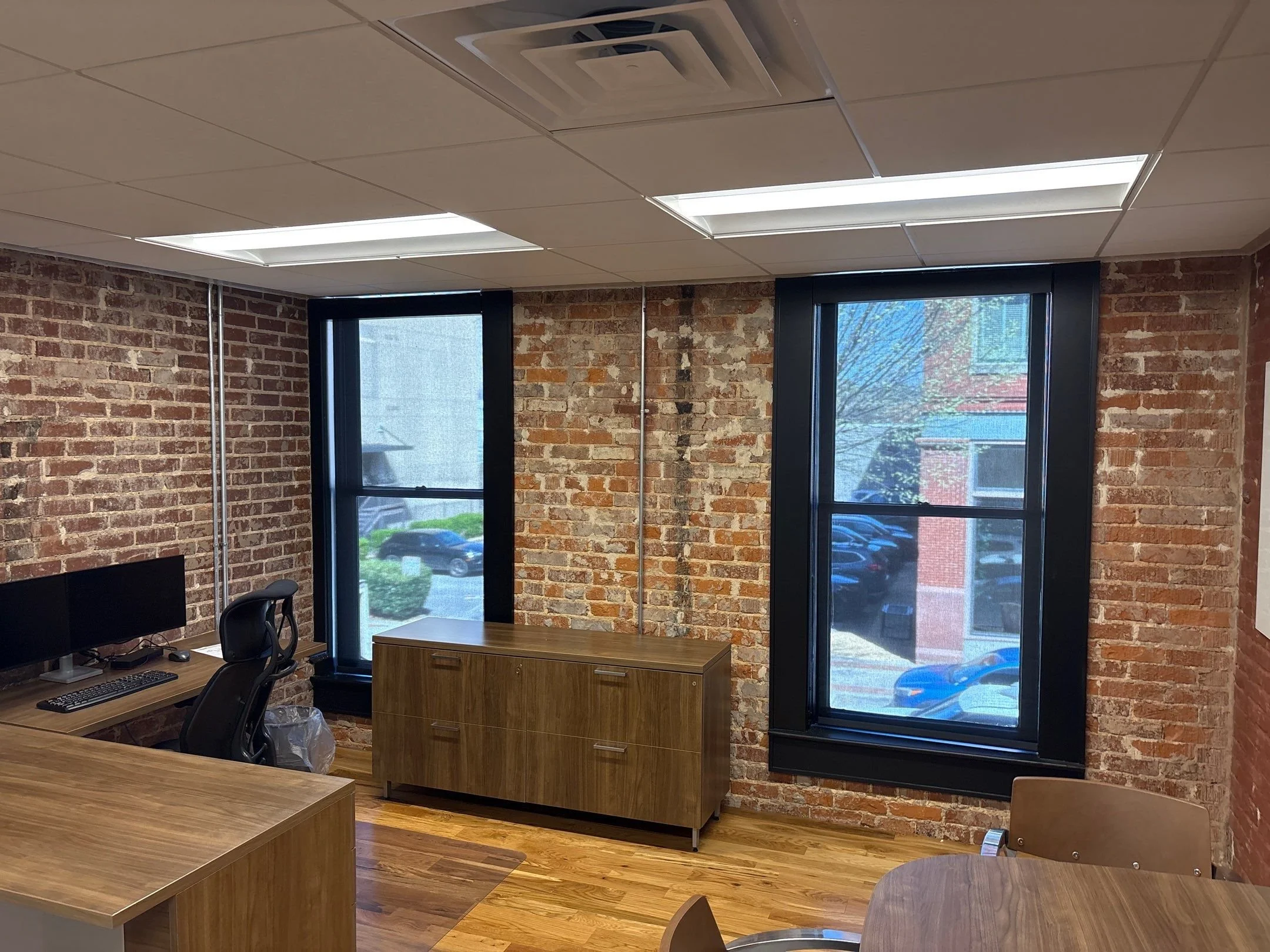 A room with exposed brick walls and large windows, black window frames, ceiling with fluorescent lights and an air conditioning vent, wooden furniture including a desk, chair, and cabinet.