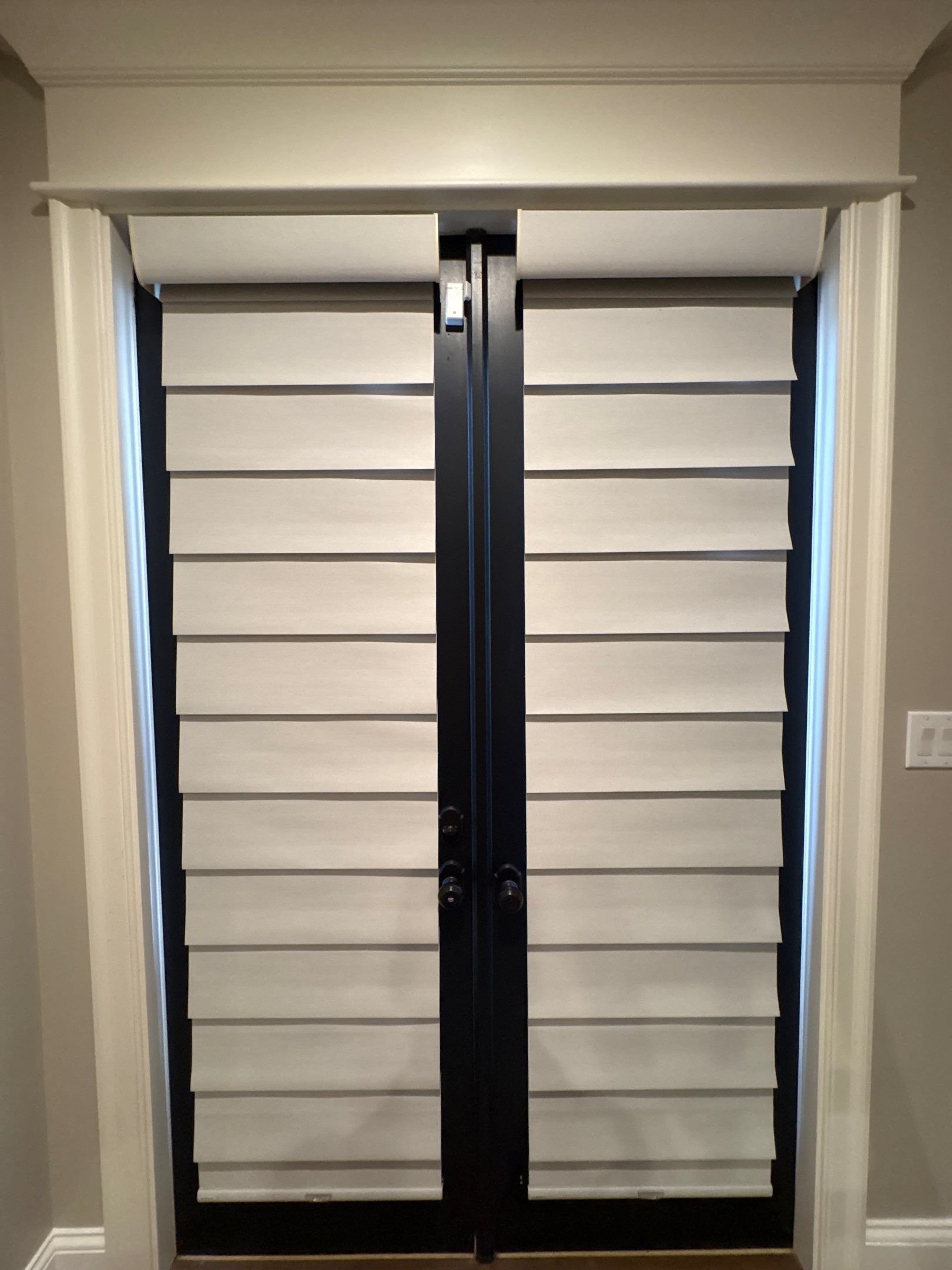 Black metal sliding door hardware with beige roller shutters inside a window frame, mounted between white trim and walls.