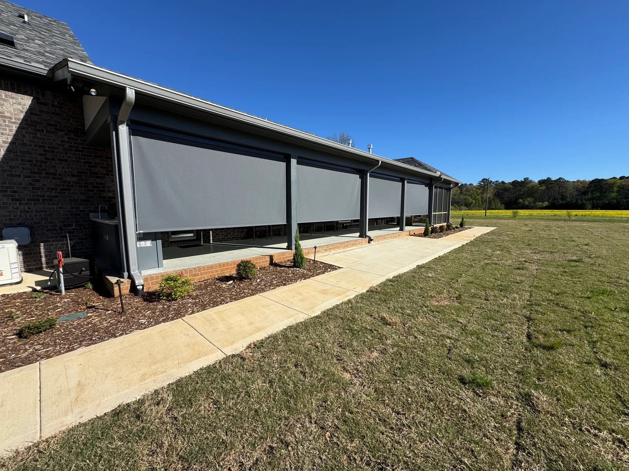 A backyard porch with a screened enclosure, small plants, a brick patio, a grassy lawn, and a clear blue sky.