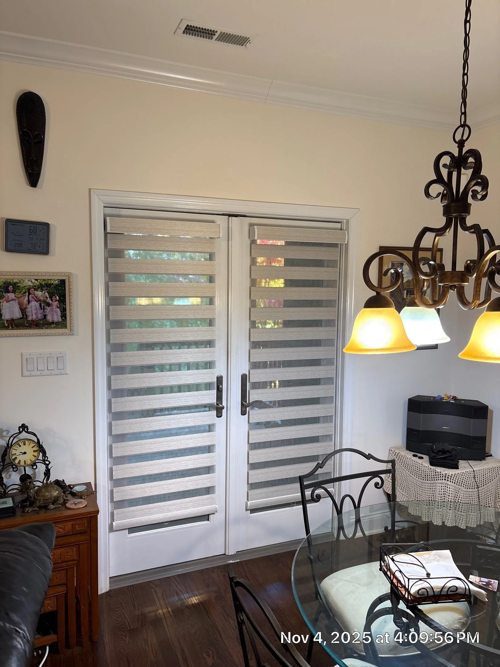 A living room corner with beige walls, white double doors with horizontal striped blinds, a chandelier, a round glass table with chairs, a clock, a framed photo of children, a black television, and a fireplace. The timestamp shows November 4, 2025, at 4:09:56 PM.