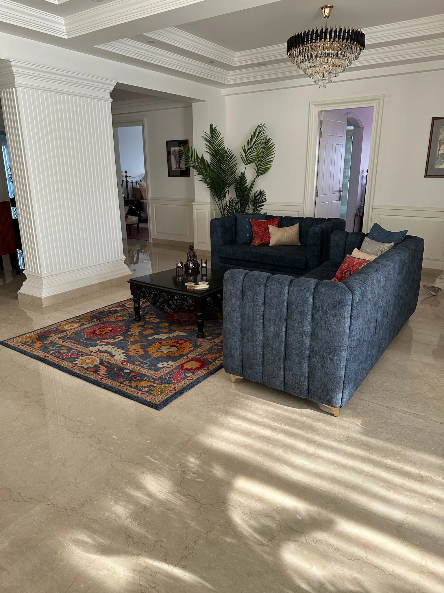 Living room with cream-colored curtains, a sliding glass door leading to a balcony with a view of trees and distant hills, a dark blue tufted sofa, a matching ottoman, and a gray sideboard.