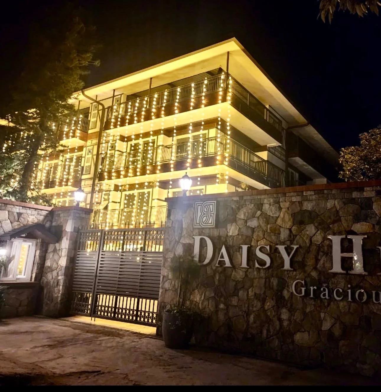 Night view of a multi-story building decorated with string lights, with a stone wall and metal gate in the foreground displaying the sign "Daisy H".