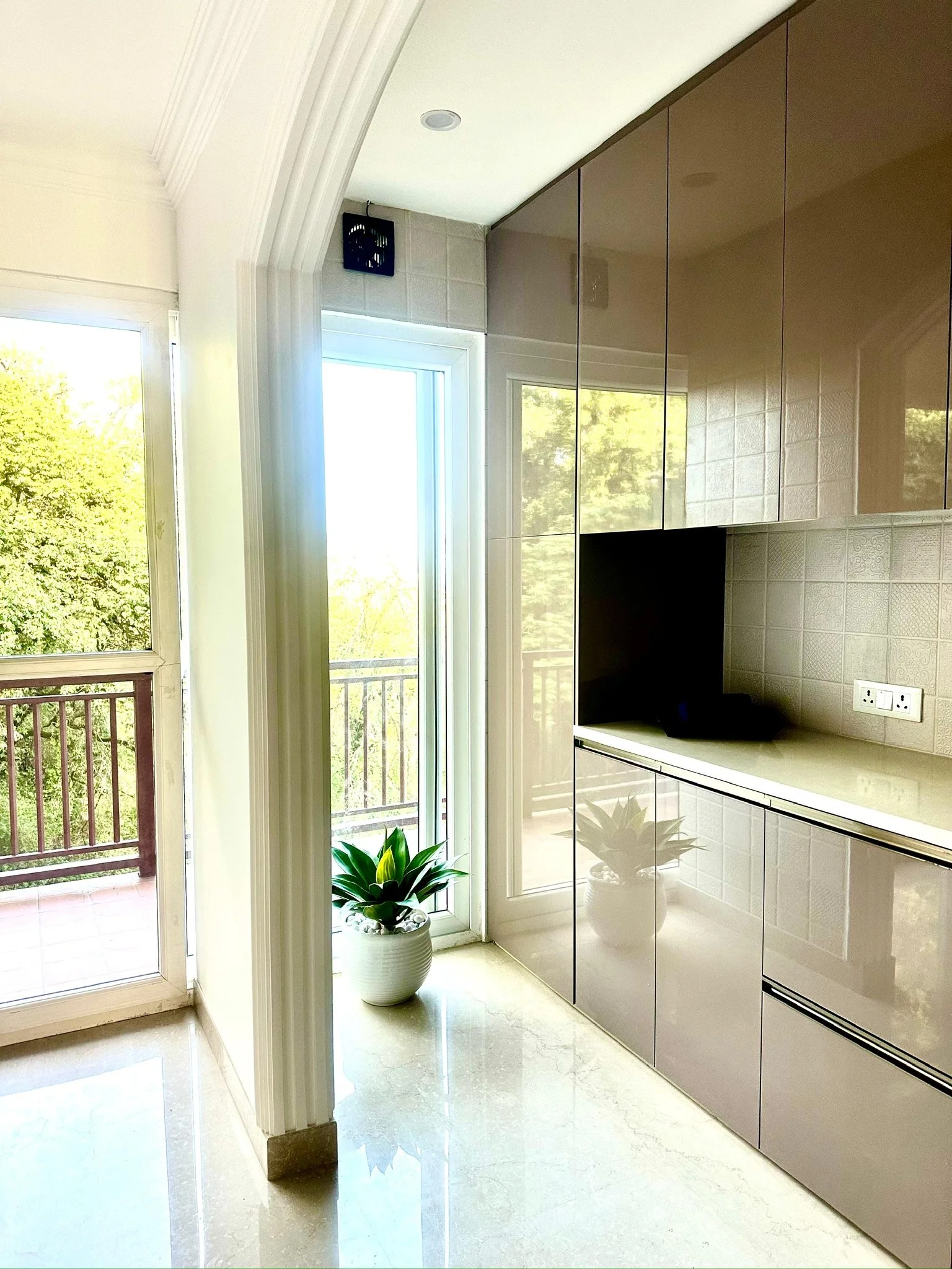 Bright kitchen corner with a potted plant, a sliding glass door leading to a balcony, and reflective beige cabinets.