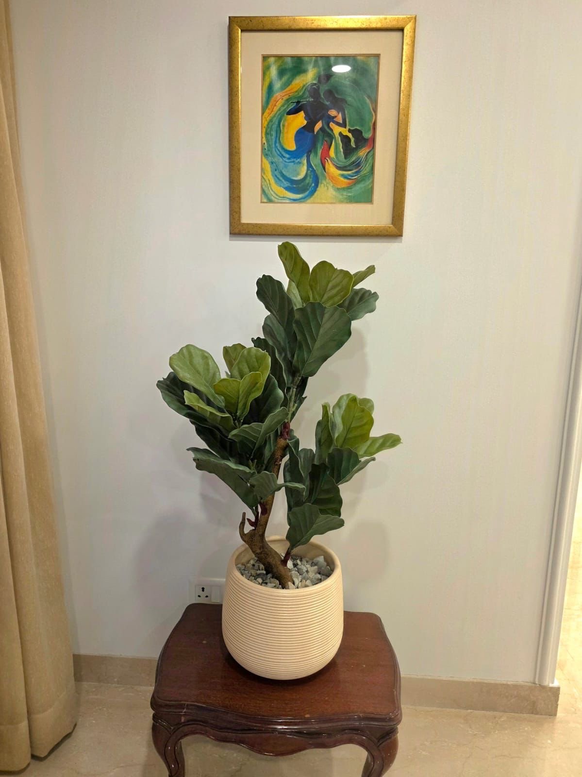 A potted green fiddle leaf fig plant on a wooden table in front of a white wall with a colorful abstract art painting hanging above it.