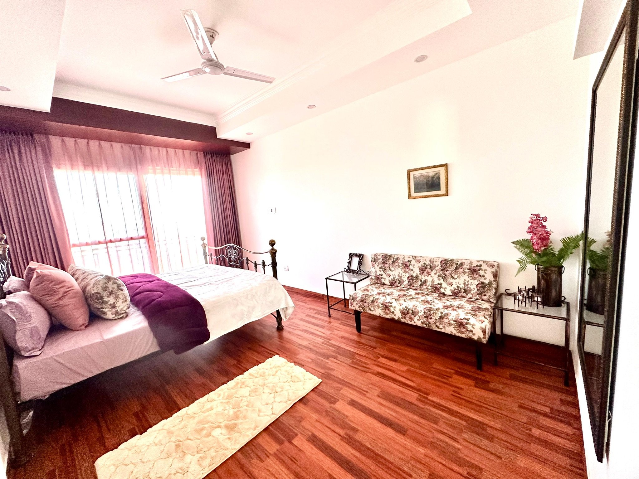 A bedroom with a metal bed frame, pink and purple bedding, a cream-colored rug, a floral-patterned sofa, two black side tables, a large window with pink curtains, a ceiling fan, and a framed picture on the wall.
